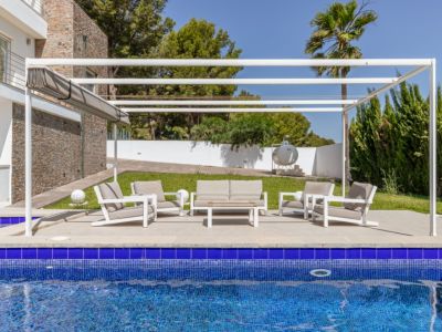Zona de estar exterior moderna con pérgola blanca y toldo retráctil, situada en una terraza pavimentada junto a la piscina de mosaico azul y jardines.