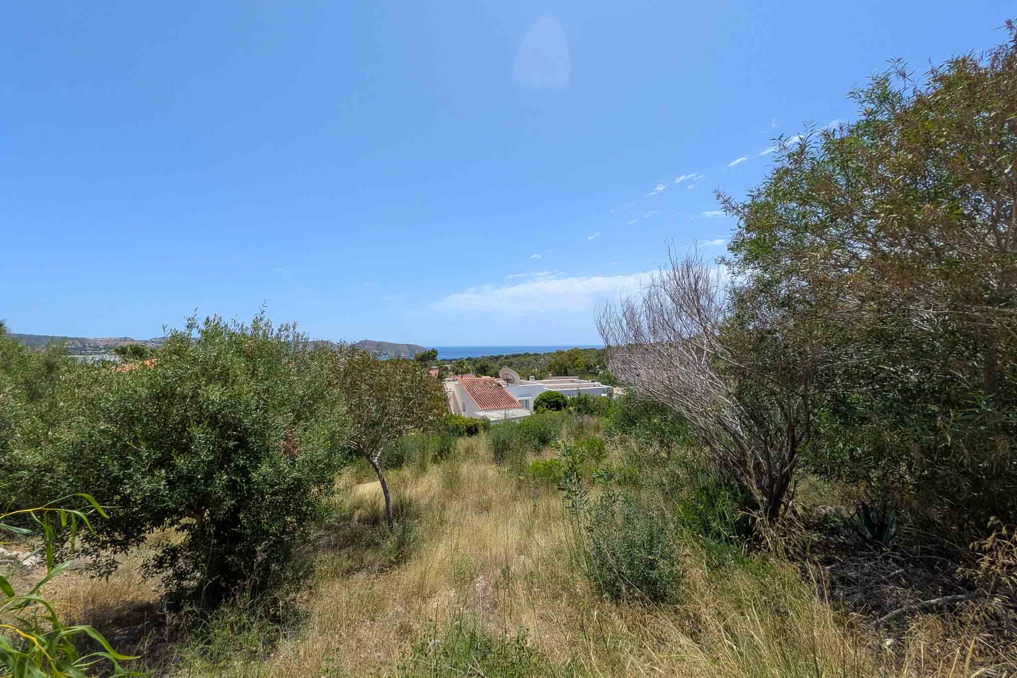 Amplia parcela con vegetación natural y vistas lejanas al mar bajo un cielo azul despejado. Techos residenciales son visibles en segundo plano, enmarcados por arbustos verdes y secos.