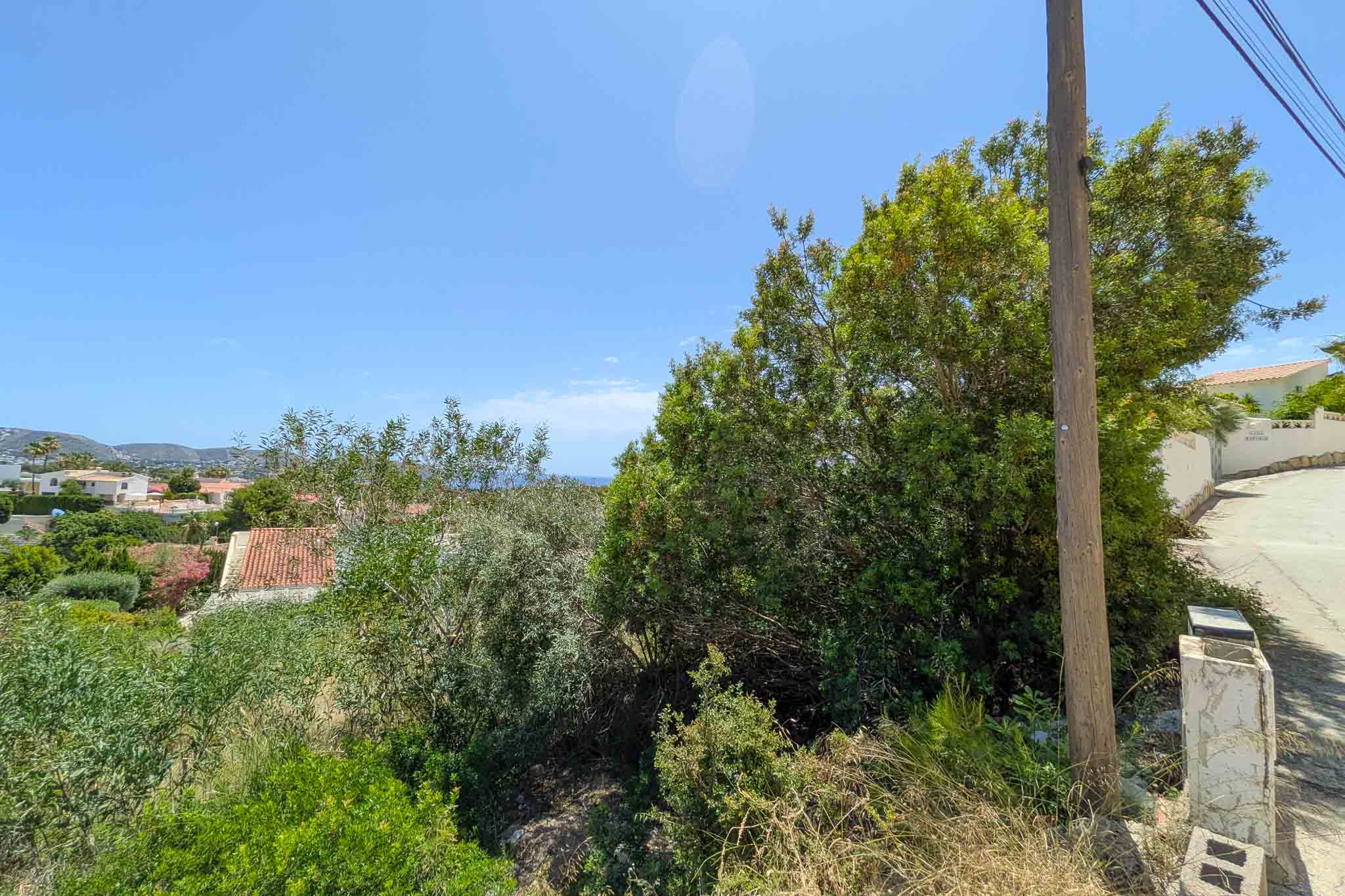 Parcela sin desarrollar con densa vegetación mediterránea, poste de servicios y camino pavimentado. Ofrece vistas lejanas a tejados residenciales, colinas y un cielo azul con atisbo de mar.