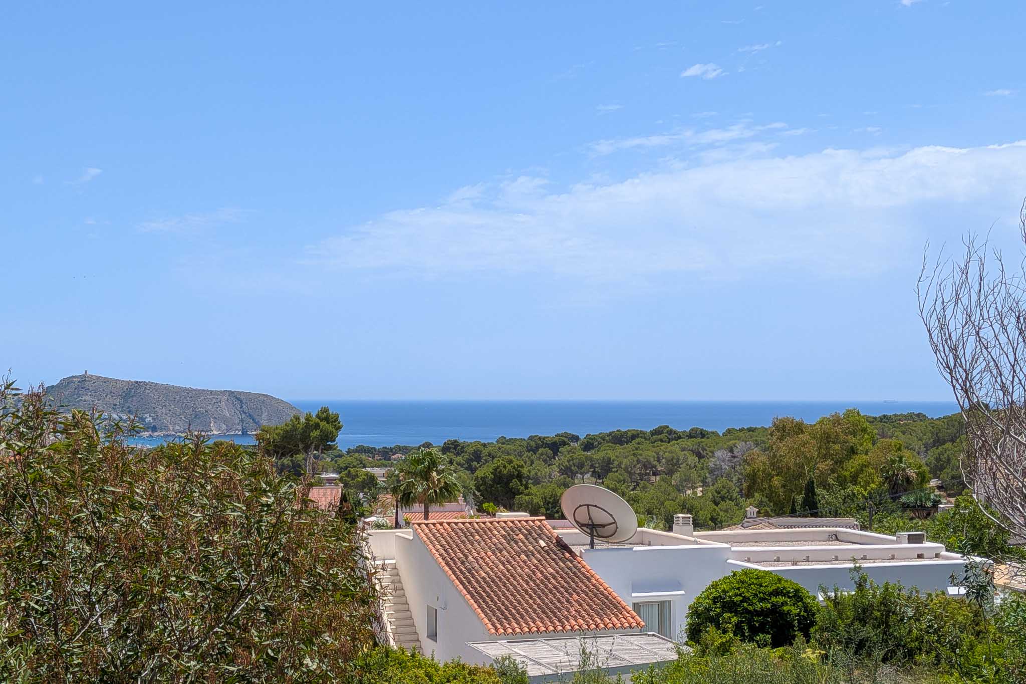 Vista elevada al mar Mediterráneo, con tejados de teja árabe y exuberante vegetación en primer plano. Un promontorio rocoso con torre se alza en la distancia bajo un cielo azul.
