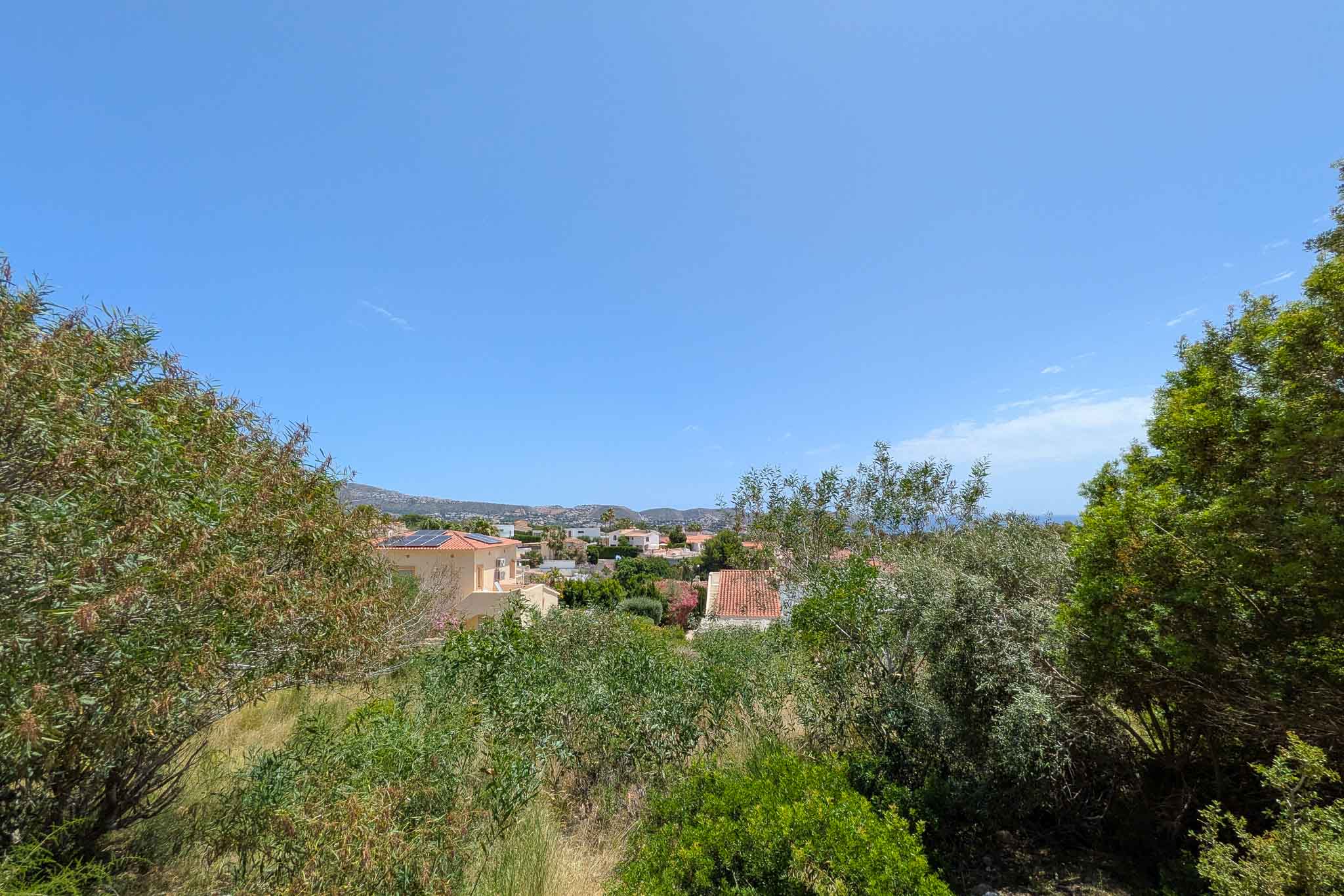 Vista panorámica desde la parcela, mostrando un entorno residencial verde con tejados tradicionales, el mar distante y las montañas bajo un cielo azul despejado.
