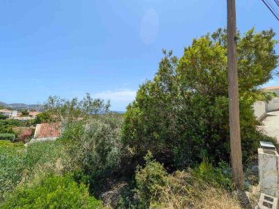 Parcela sin desarrollar con densa vegetación mediterránea, poste de servicios y camino pavimentado. Ofrece vistas lejanas a tejados residenciales, colinas y un cielo azul con atisbo de mar.