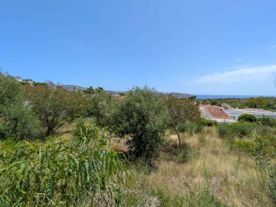 Parcela sin urbanizar con vegetación mediterránea natural, ofreciendo vistas parciales al mar enmarcadas por tejados residenciales y colinas lejanas bajo un cielo azul claro.