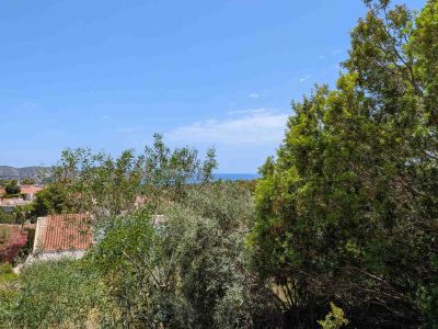 Vista panorámica desde la parcela, con exuberante vegetación mediterránea en primer plano, tejados residenciales y el mar Mediterráneo en el horizonte bajo un cielo azul despejado.