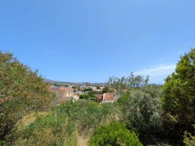 Vista panorámica desde la parcela, mostrando un entorno residencial verde con tejados tradicionales, el mar distante y las montañas bajo un cielo azul despejado.