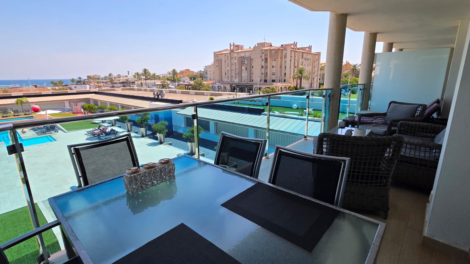 Amplia terraza con mesa de comedor de cristal y zona de estar de mimbre, ofreciendo vistas a la piscina comunitaria y al Mediterráneo bajo un cielo despejado.