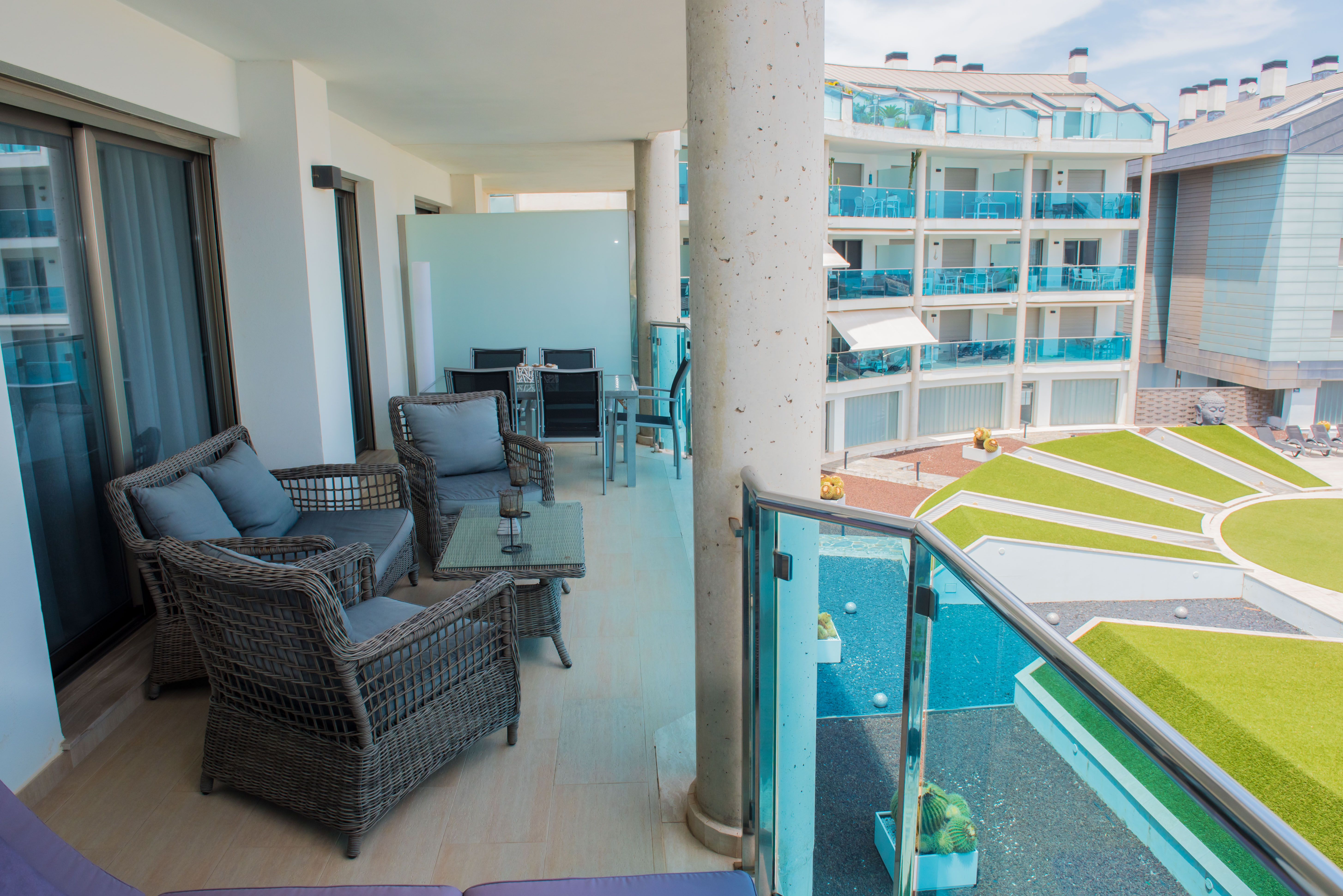 Amplia terraza cubierta con mobiliario de ratán, mesa de centro de cristal y suelo de baldosas claras. Vistas a jardines comunitarios modernos y edificio adyacente con balcones de cristal azul.