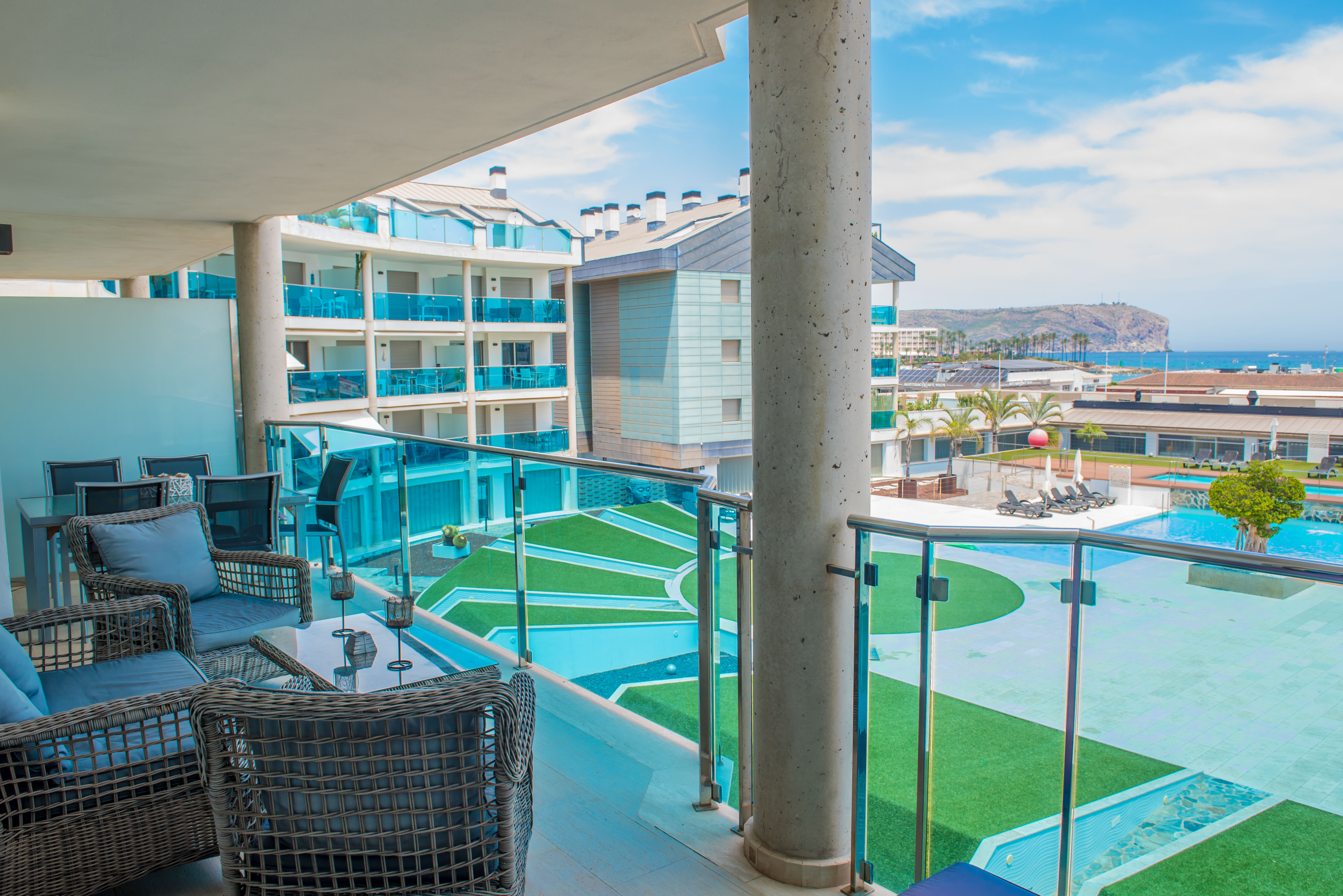 Amplia terraza privada con mobiliario de exterior de ratán y vistas directas a las piscinas comunitarias y al mar. Barandilla de cristal y suelo de gres claro.