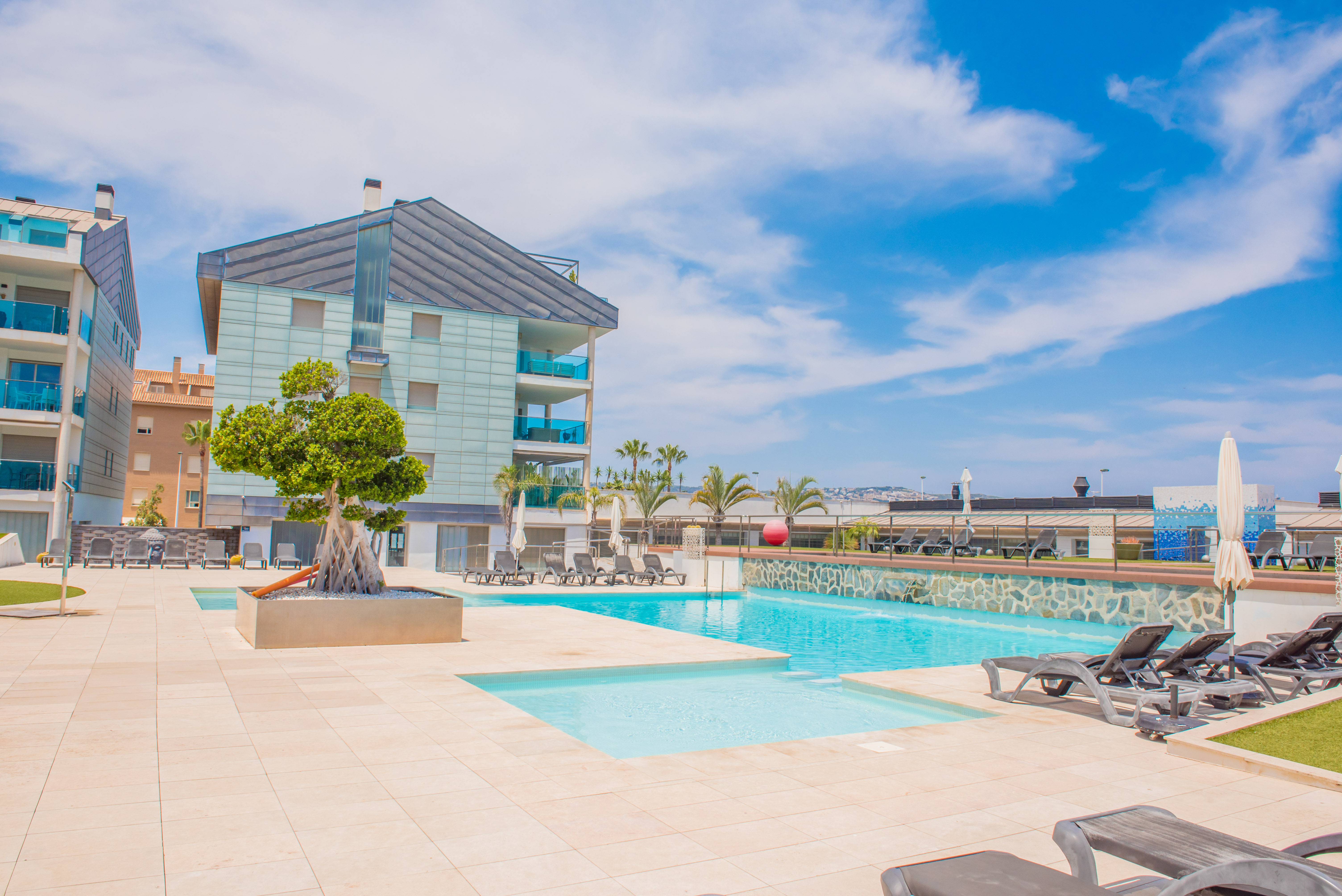 Amplia zona de piscina comunitaria con dos piscinas, pavimentación clara y numerosas tumbonas. Edificios residenciales modernos con balcones de cristal azulado enmarcan este espacio soleado.