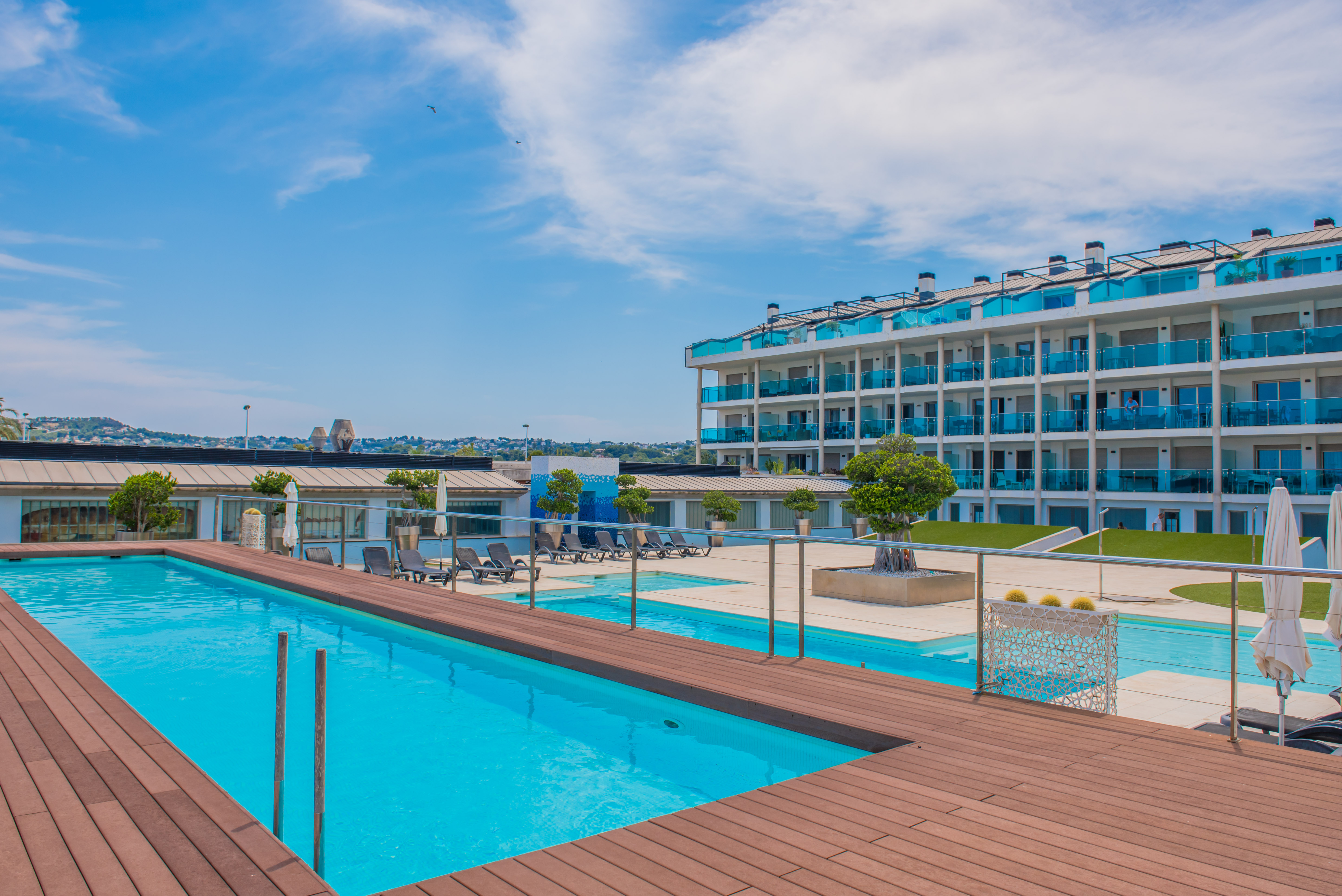 Amplia zona de piscina comunitaria con una piscina de azul brillante y una tarima de madera cálida. El moderno edificio de apartamentos, con balcones de cristal azul, domina este espacio exterior sereno.