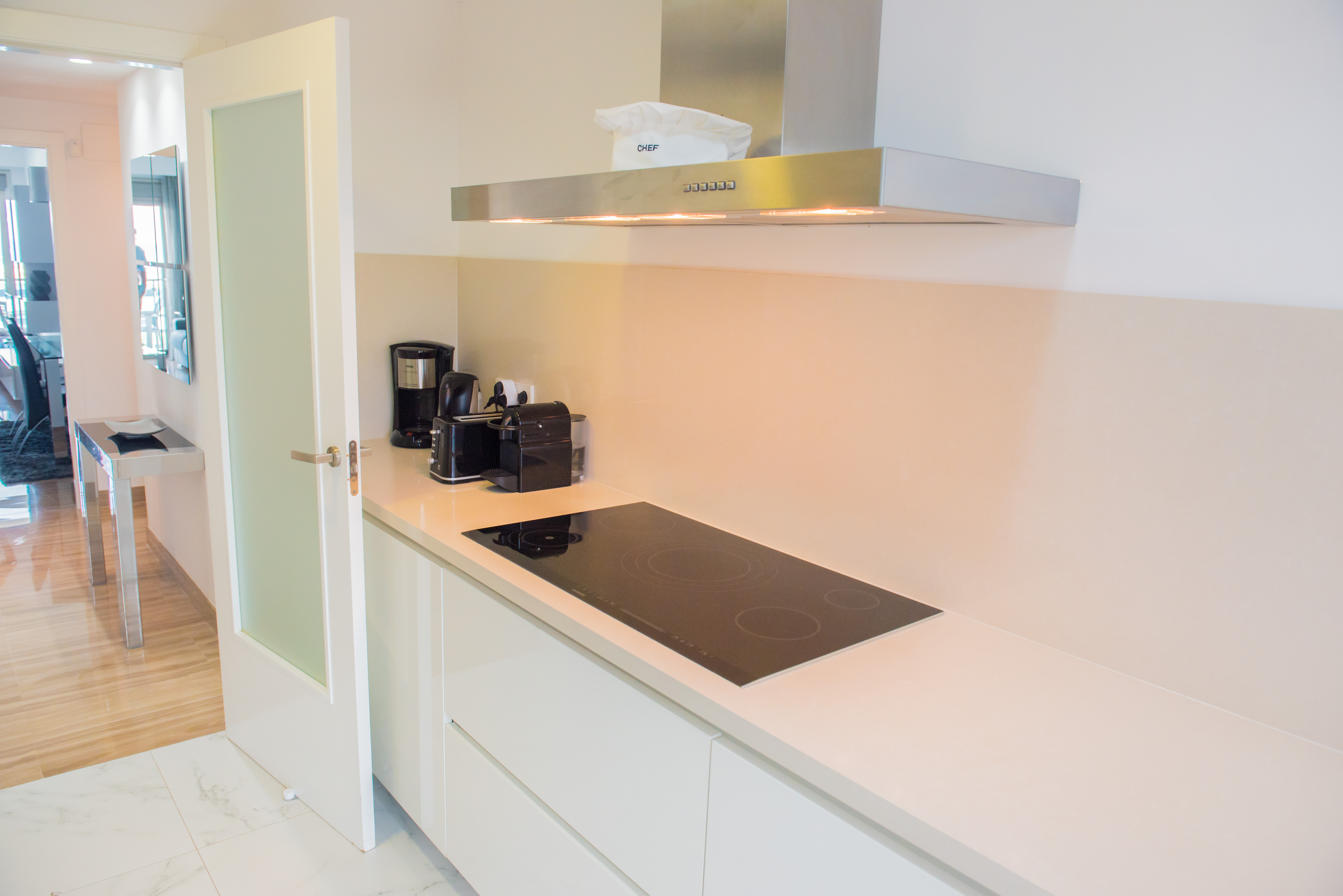 Cocina moderna con encimera clara, placa de inducción negra y campana extractora de acero inoxidable. Suelos de mármol y acceso a una sala con parquet.