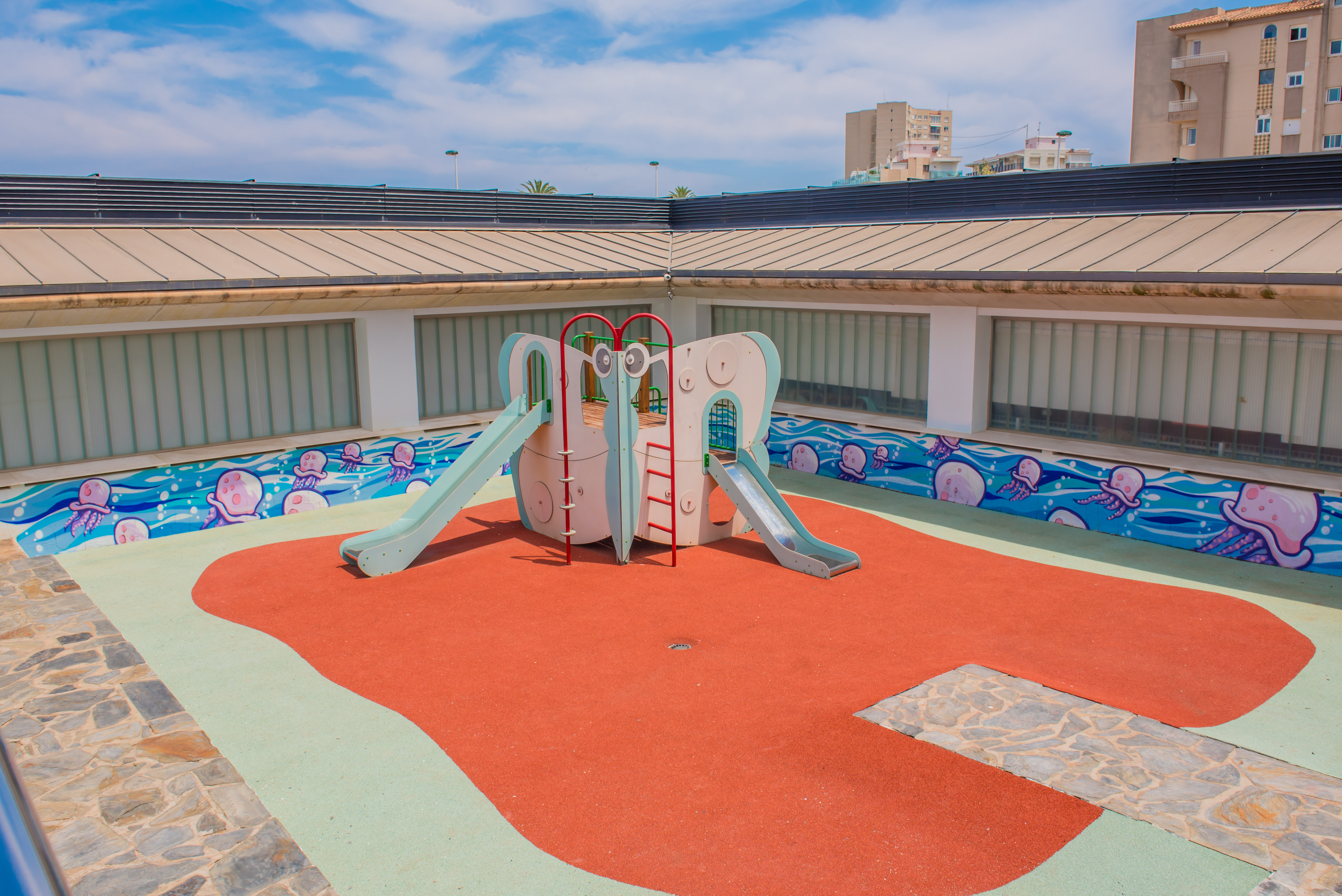 Colorida zona de juegos infantiles con estructura central de toboganes sobre superficie de caucho rojo y verde. Un vibrante mural de medusas decora la pared, bajo paneles de vidrio esmerilado.