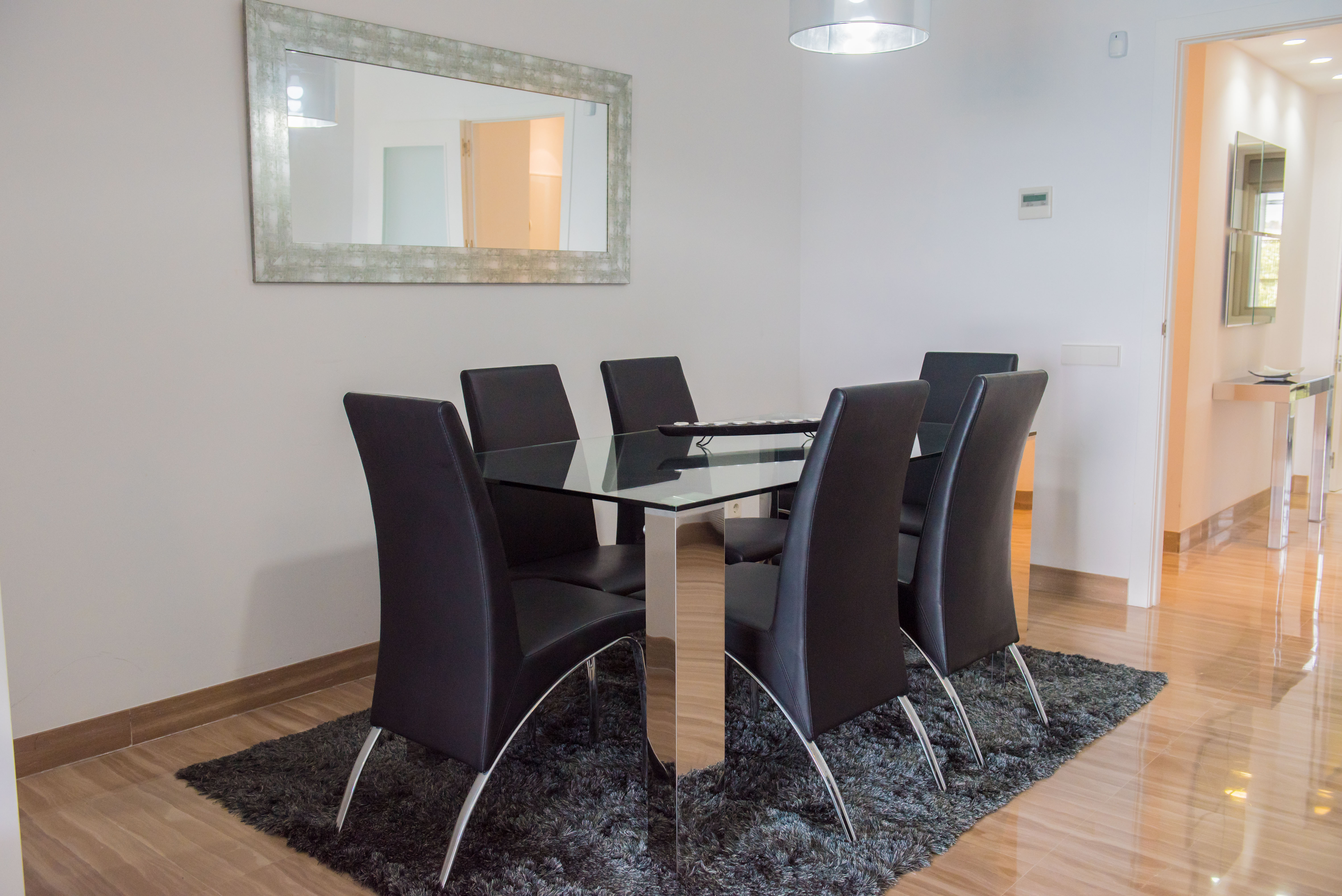 Comedor moderno con mesa de cristal y seis sillas de polipiel negra. Destaca el espejo de marco envejecido y el suelo de mármol pulido.