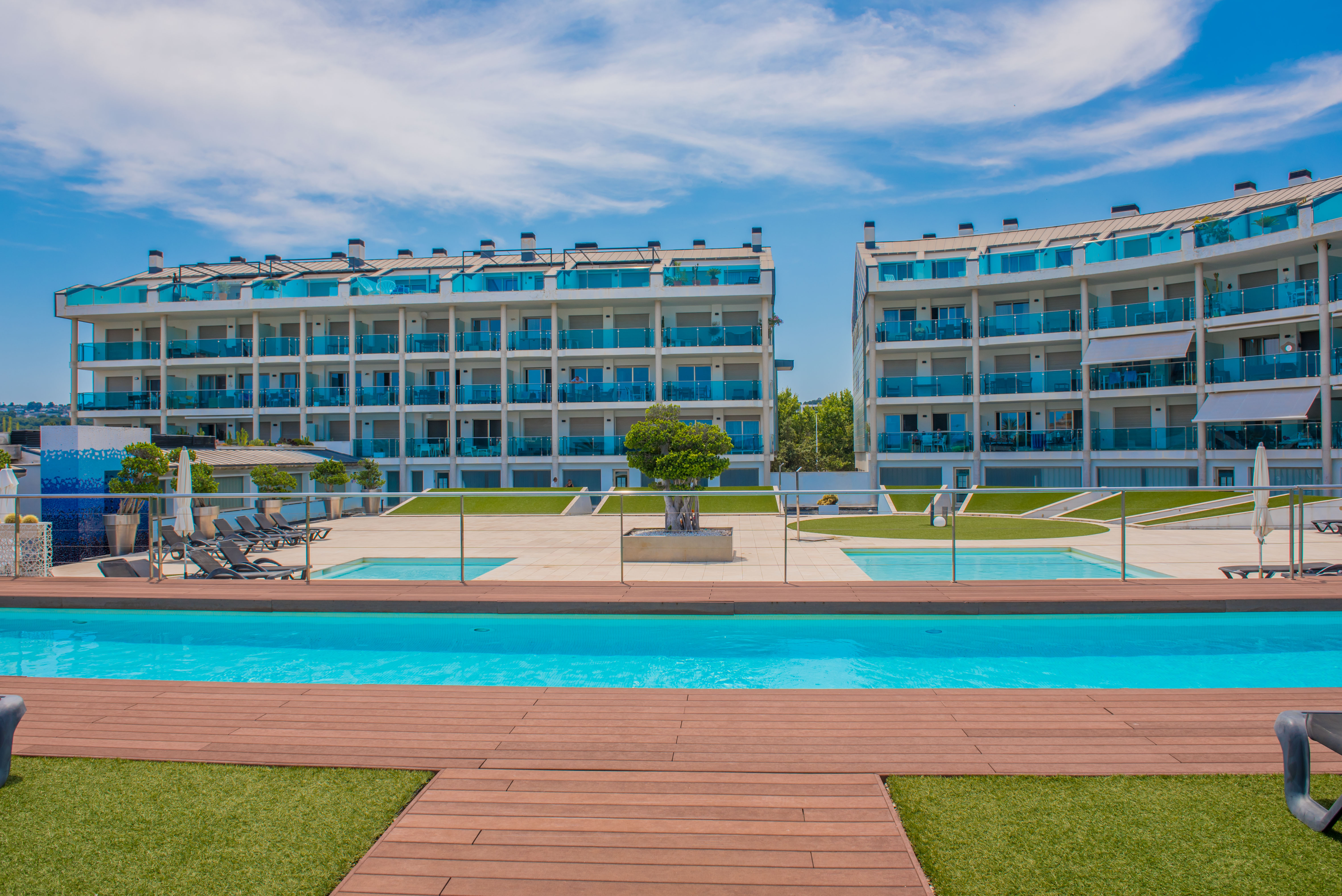 Complejo residencial moderno con dos edificios de balcones curvos de cristal azul, rodeando una gran piscina comunitaria con elegante tarima de madera y césped artificial.