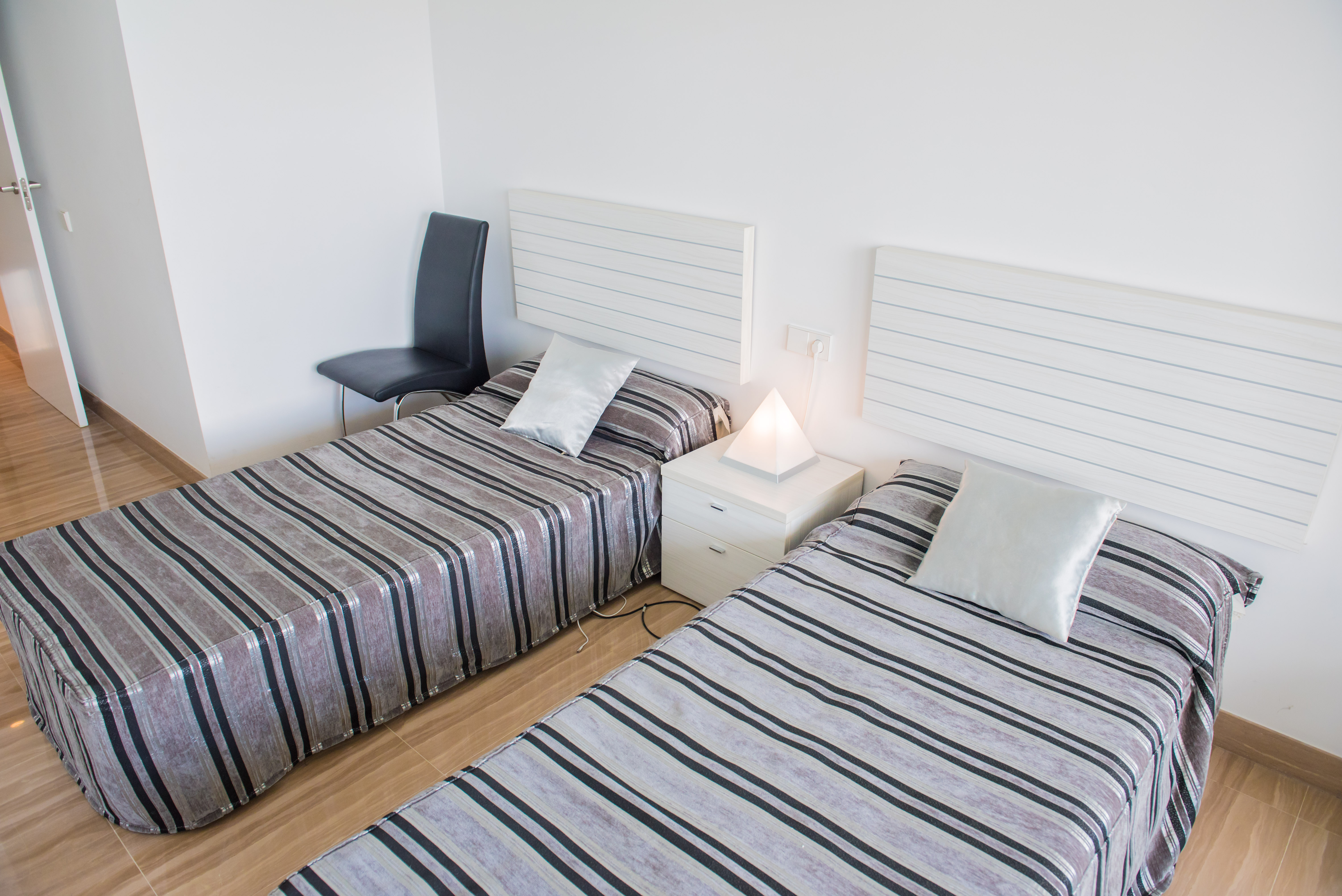 Dormitorio con dos camas individuales, cabeceros blancos de lamas y ropa de cama a rayas. Una lámpara piramidal ilumina la mesita de noche entre ellas.
