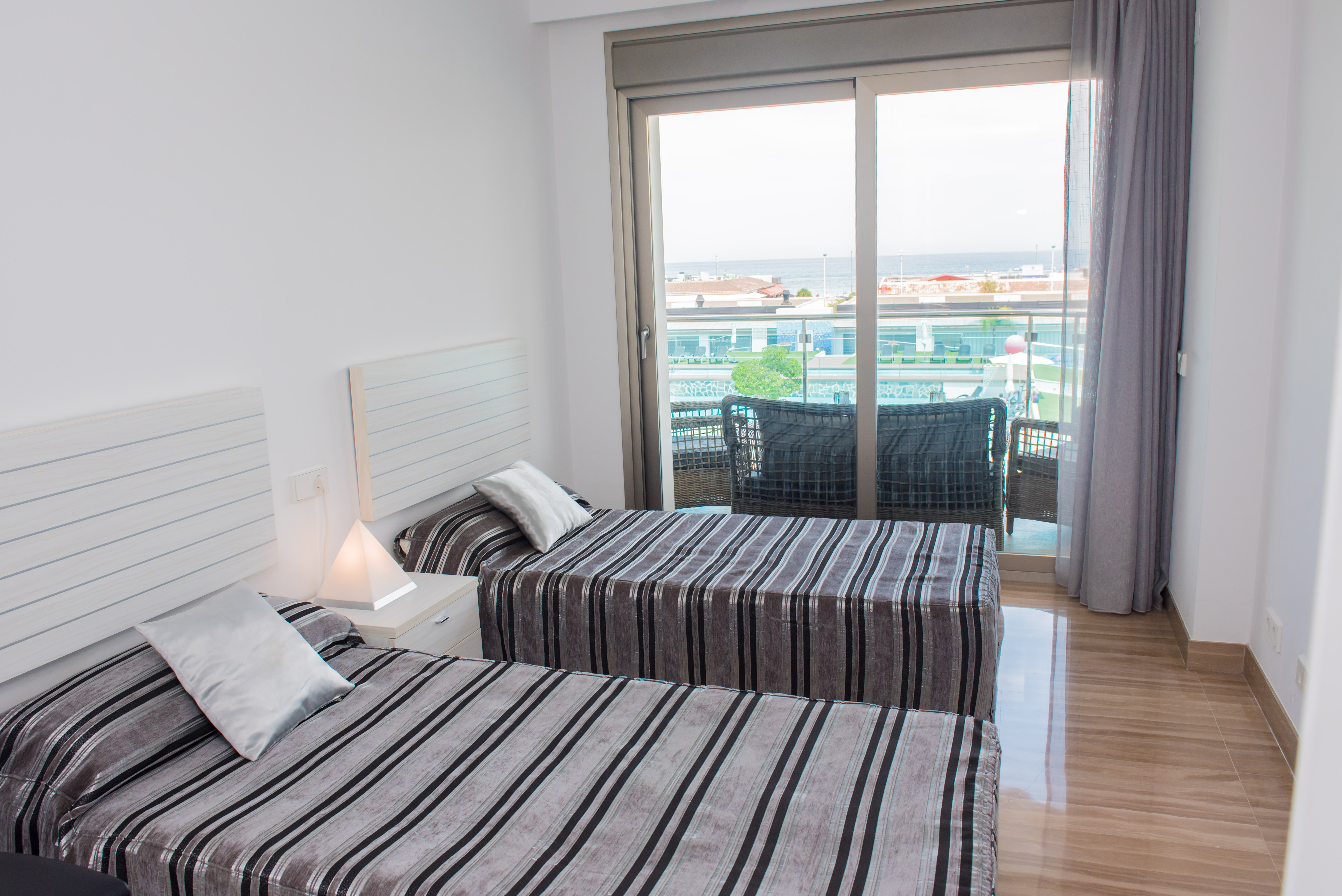 Dormitorio luminoso con dos camas individuales, ropa de cama a rayas y mesita de noche. Puertas correderas dan a un balcón con asientos exteriores y vistas parciales al mar. Suelo pulido.