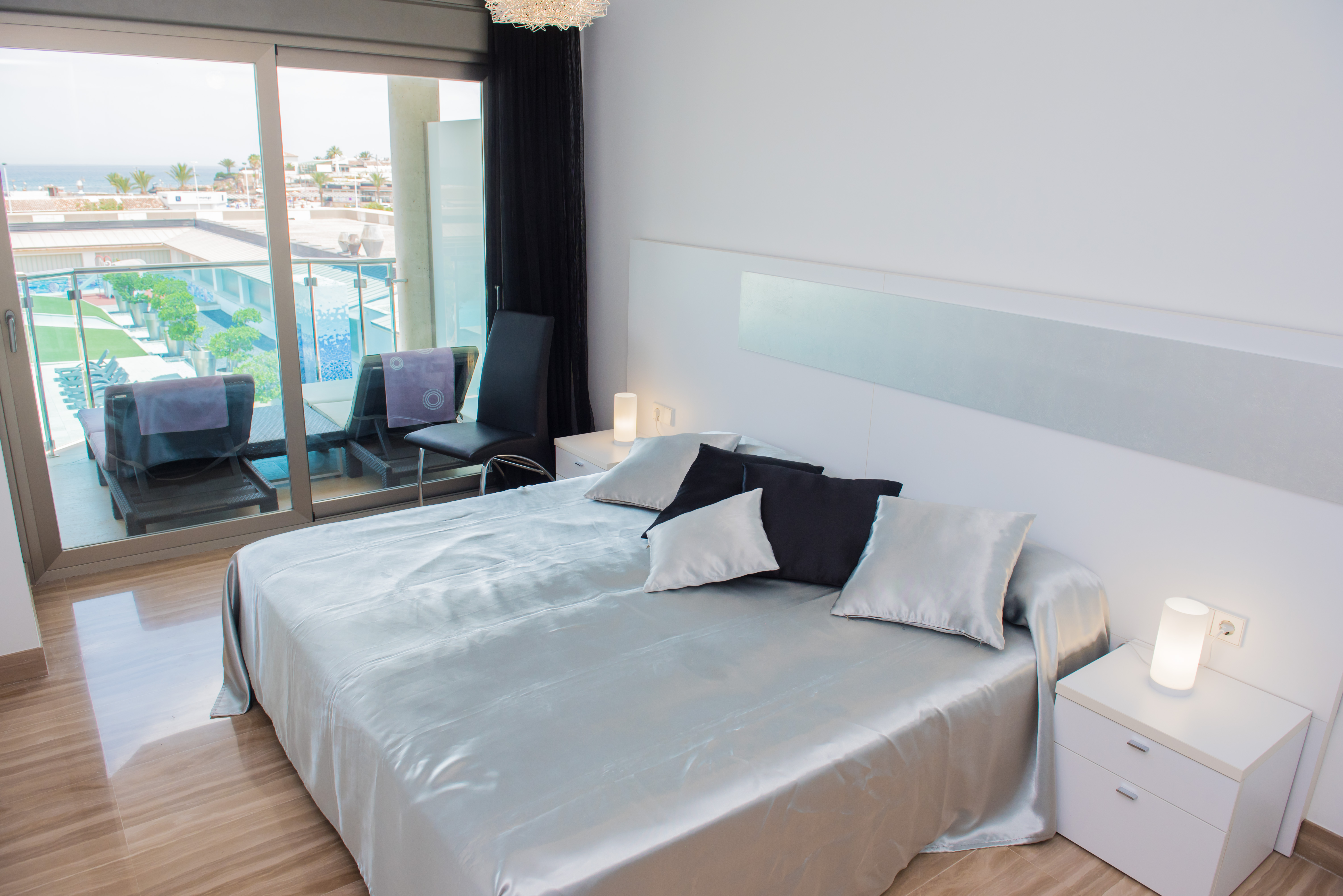 Dormitorio principal moderno con cama grande, ropa de cama satinada plateada, suelos de gres brillante y puertas correderas a la terraza con vistas al mar y piscina.