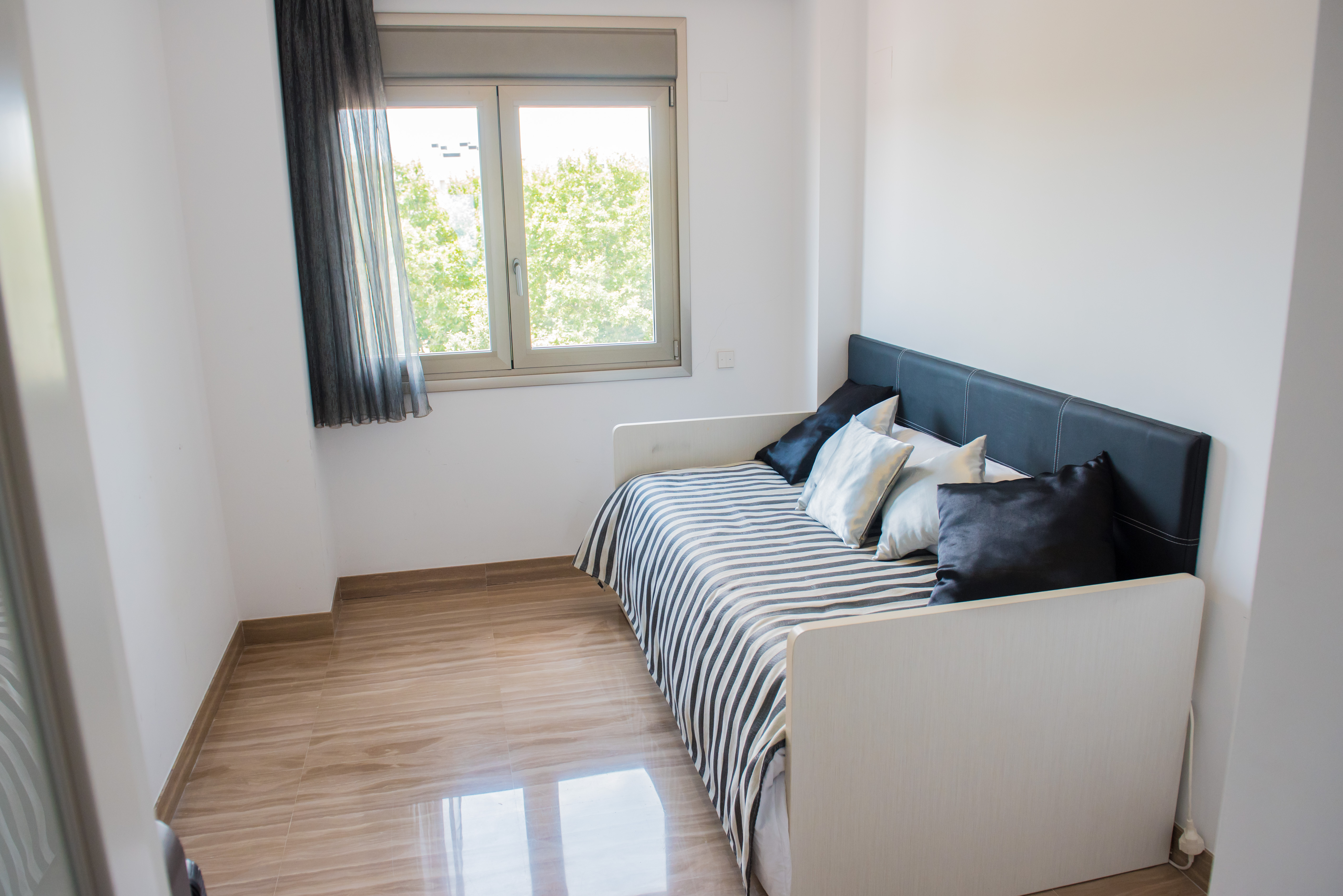 Habitación individual con cama nido, cabecero tapizado negro y ropa de cama a rayas. El suelo de mármol claro pulido y la ventana ofrecen vistas a la vegetación.
