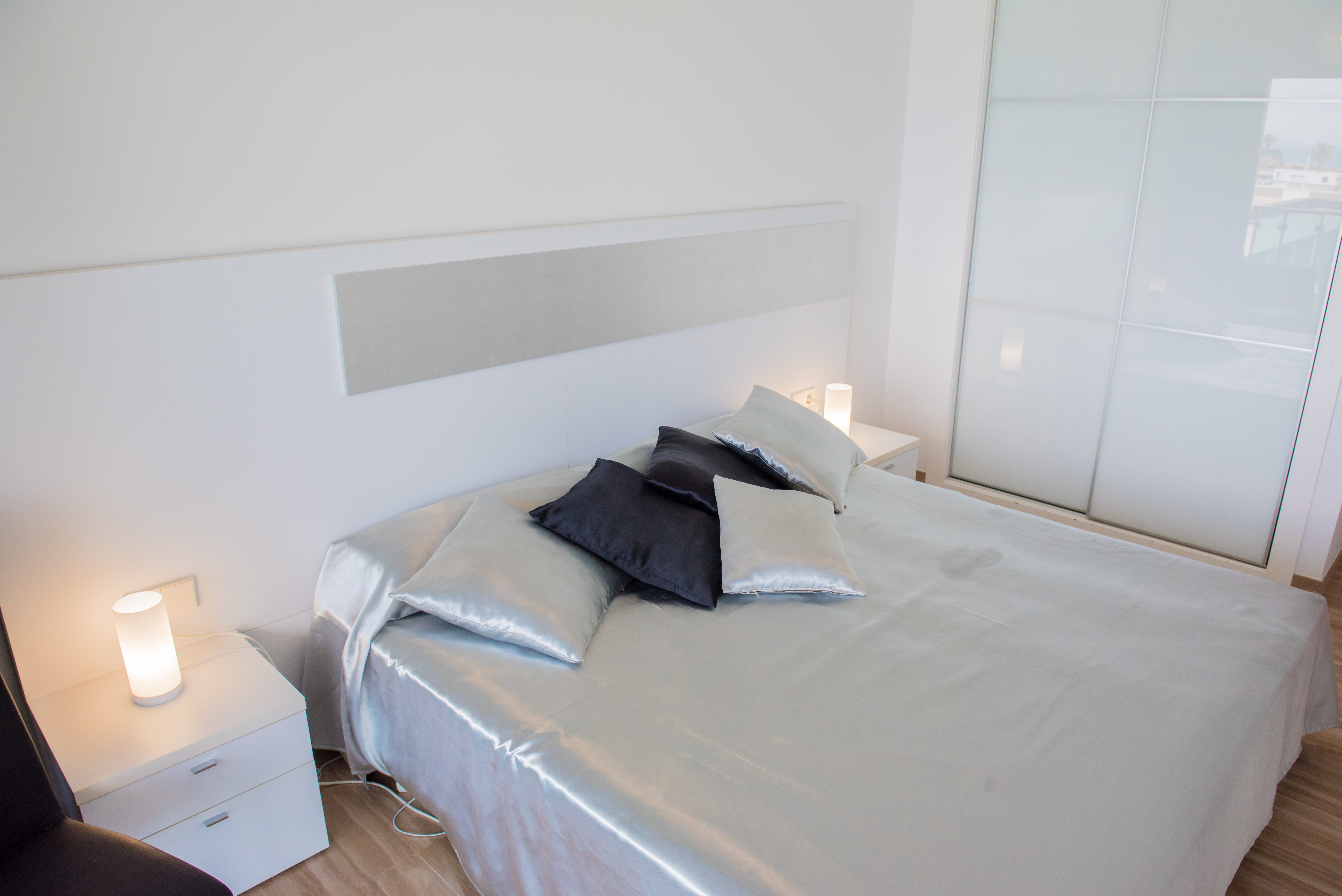 Habitación moderna con cama de sábanas satinadas, cabecero blanco y mesitas de noche con lámparas cilíndricas. Armario empotrado blanco con puertas correderas de cristal esmerilado.