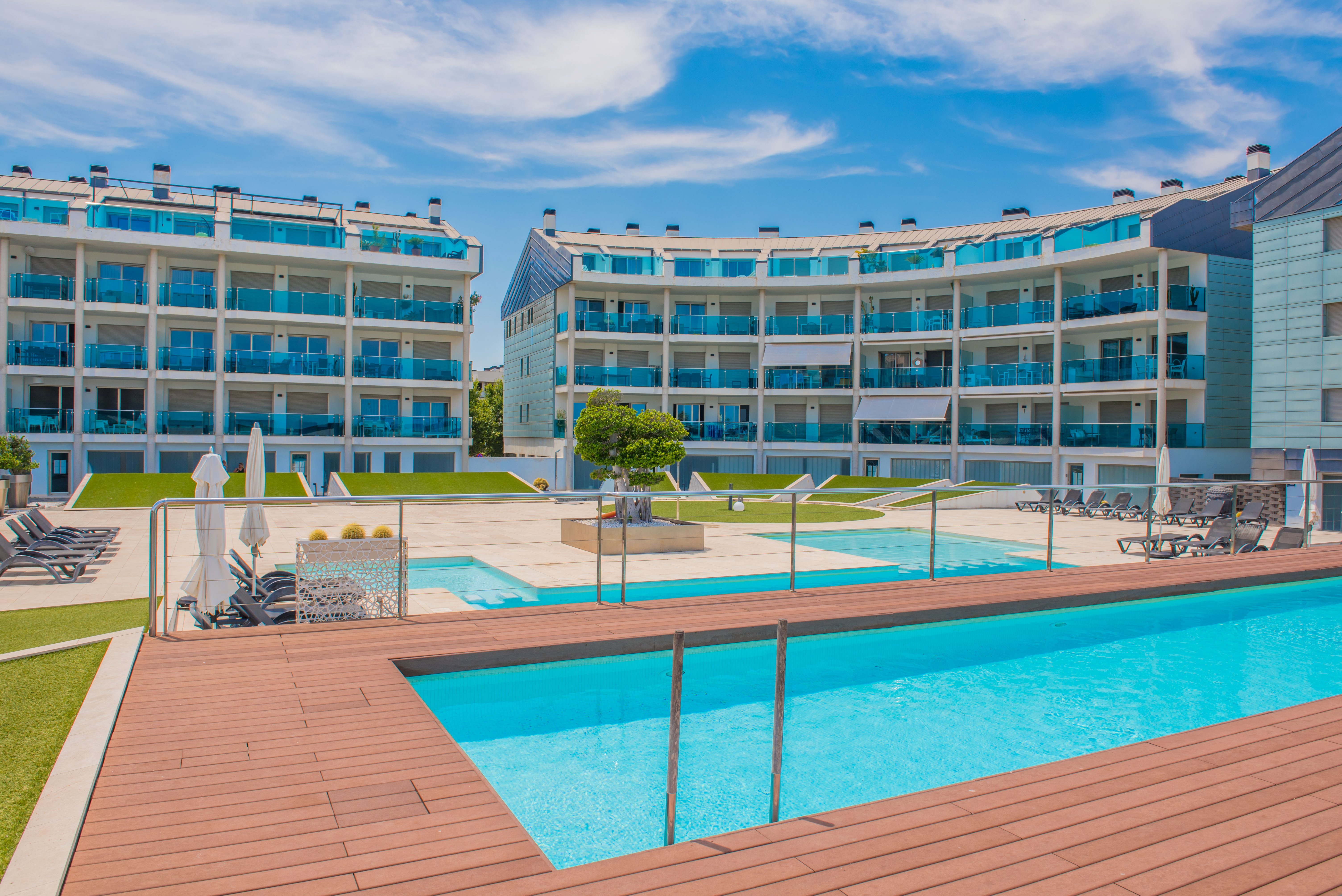 La zona de piscina comunitaria presenta dos piscinas de agua azul cristalina, rodeadas de tarima de madera, tumbonas y césped artificial, frente a edificios de apartamentos modernos.