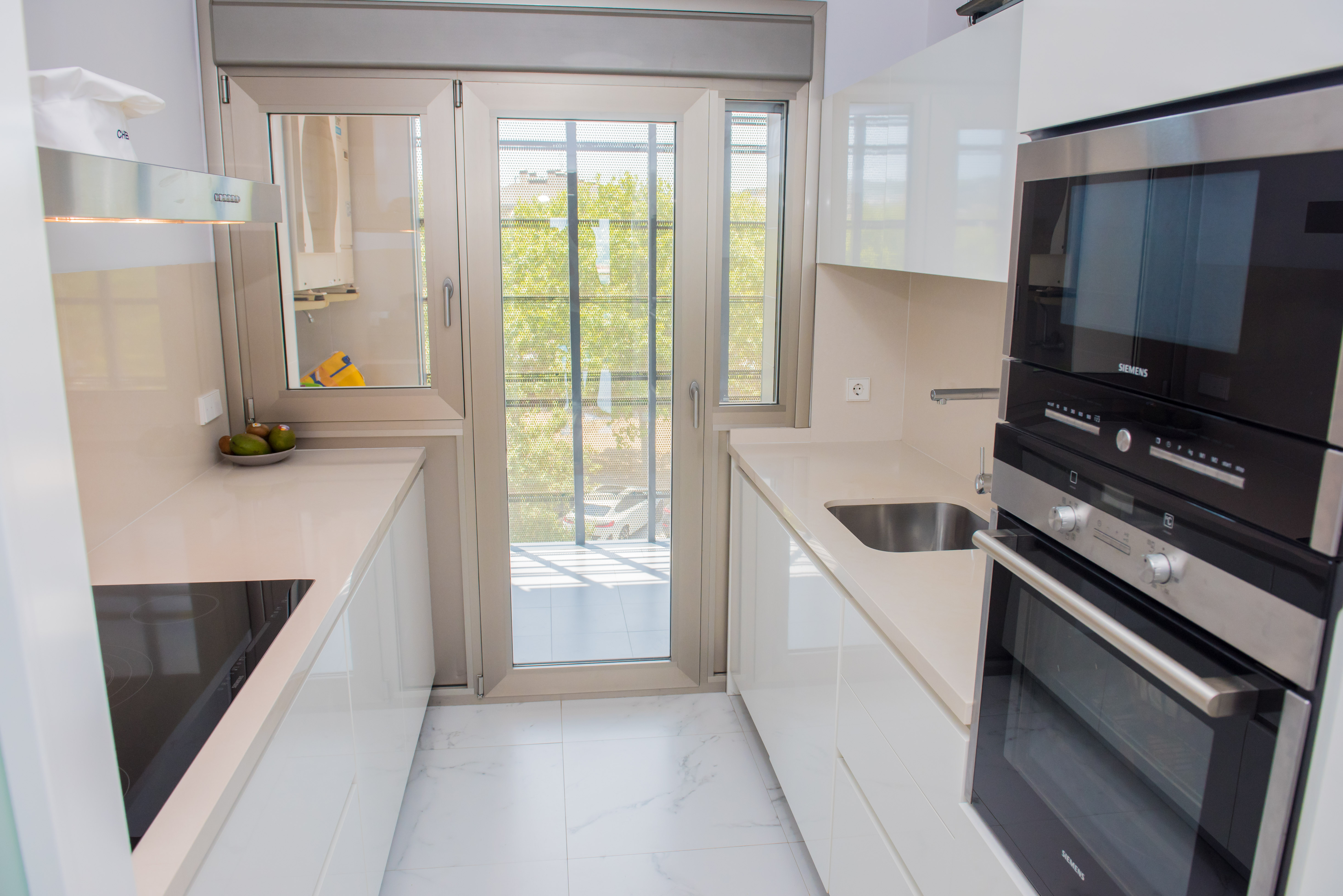 Moderna cocina tipo galera con elegantes muebles blancos, encimeras claras y electrodomésticos Siemens integrados. La puerta doble acristalada proporciona luz natural y acceso a un balcón.