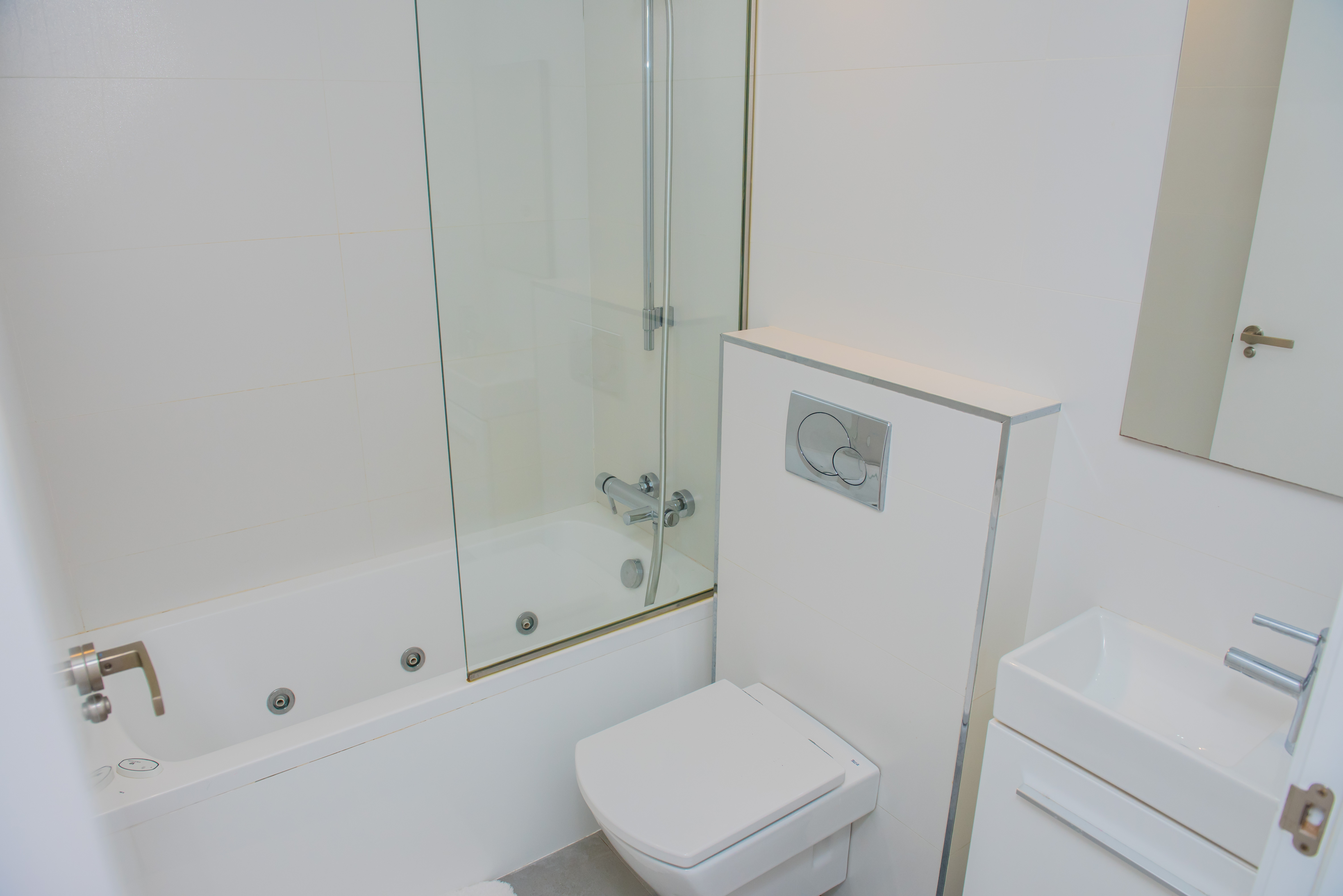 Moderno cuarto de baño blanco con bañera de hidromasaje, mampara de cristal, inodoro suspendido y lavabo rectangular. Azulejos blancos y grifería cromada.