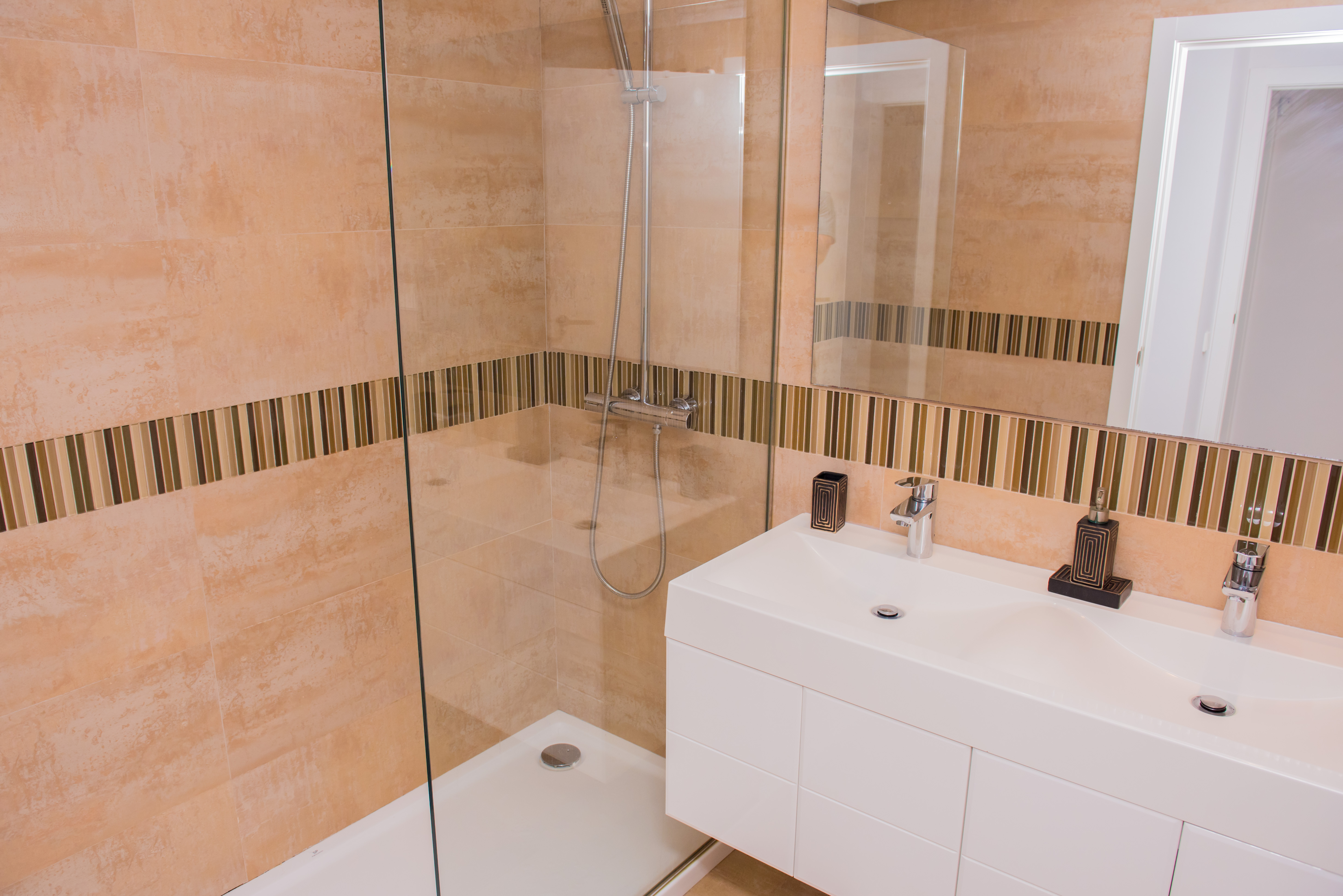 Moderno cuarto de baño con ducha a ras de suelo y mampara de cristal. Destaca el lavabo doble y los azulejos beige con franja decorativa de mosaico.