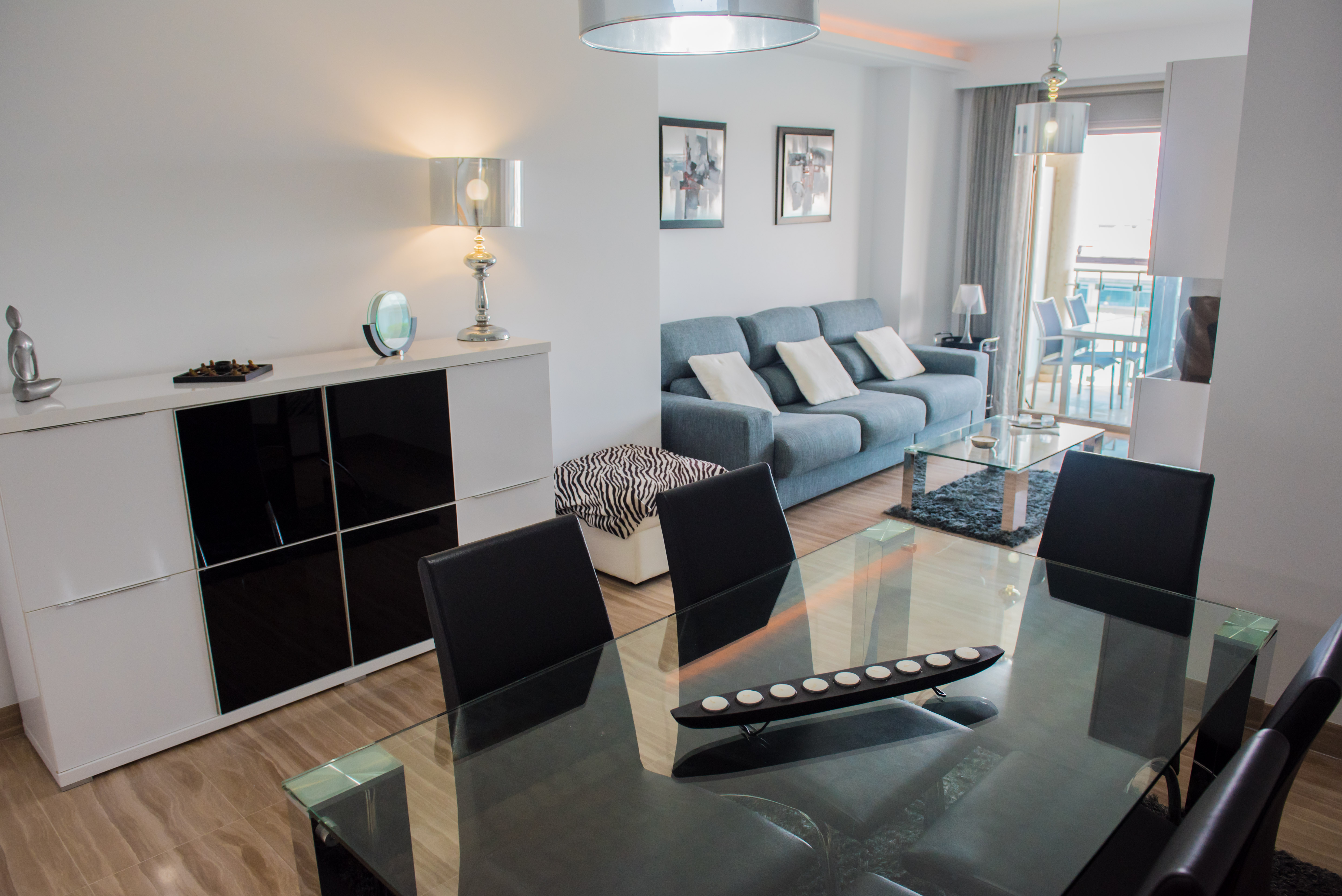 Salón-comedor de concepto abierto con mesa de cristal, sillas negras y sofá azul. El suelo de baldosas y un aparador blanco moderno complementan el espacio, con acceso a la terraza.