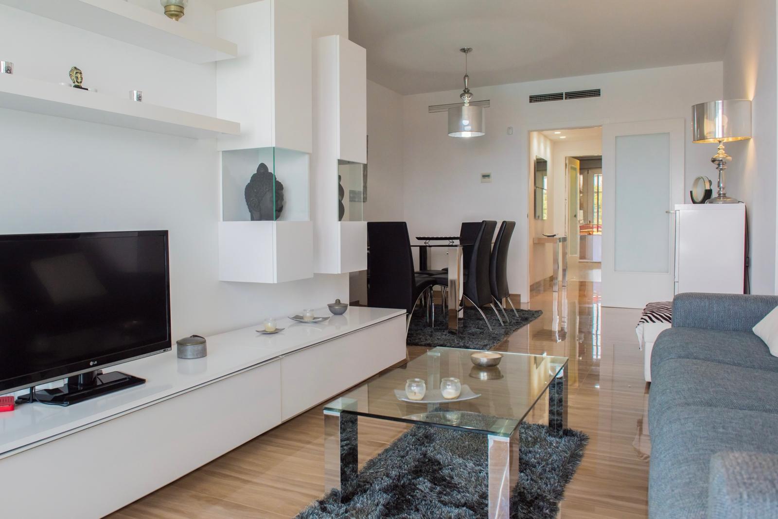 Salón-comedor moderno con mueble modular blanco, mesa de centro de cristal sobre alfombra oscura y conjunto de comedor con sillas negras. Suelos pulidos y aire acondicionado integrado.