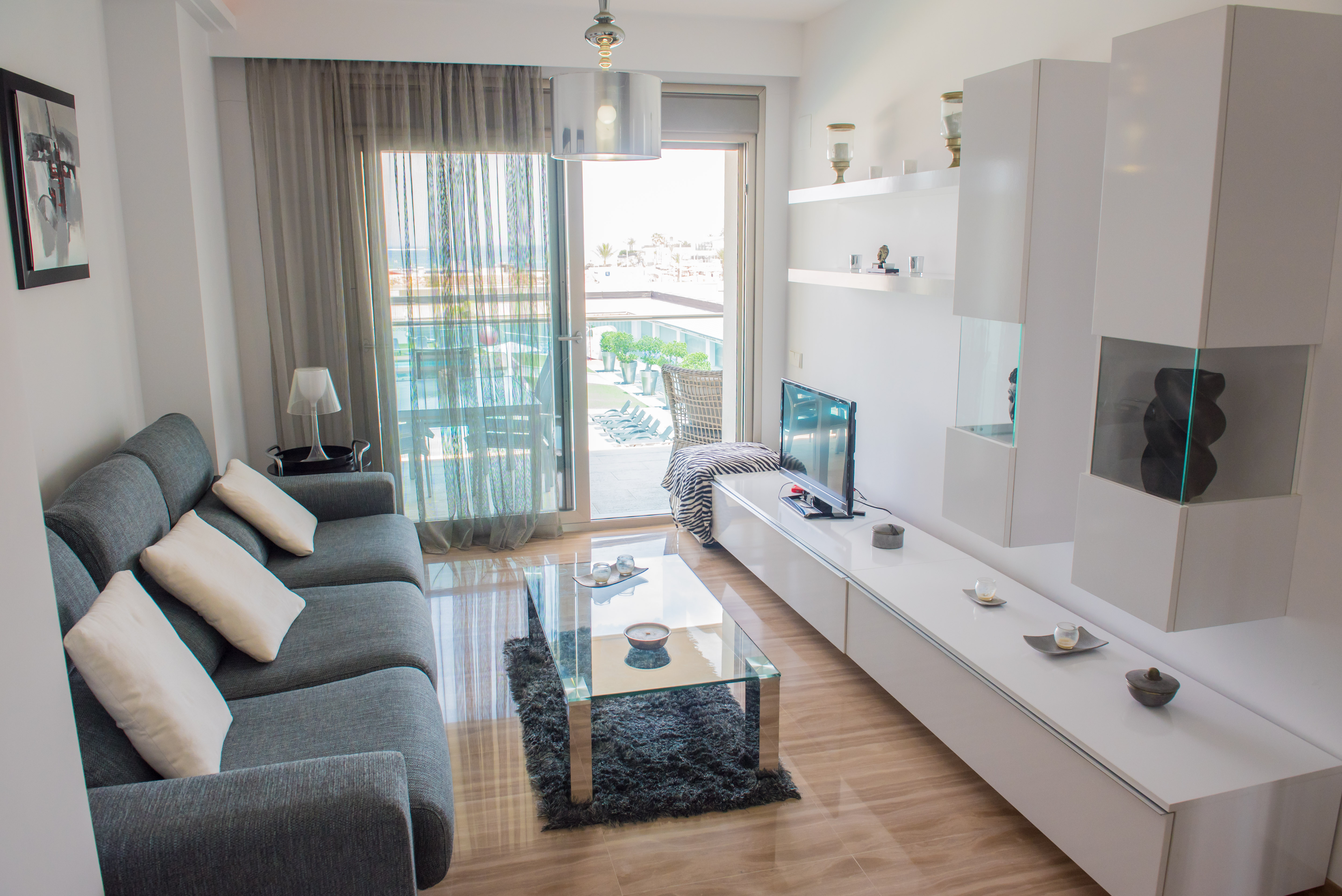 Salón moderno con sofá seccional gris y mesa de centro de cristal. Puertas correderas de vidrio dan a una terraza con vistas a la piscina y al mar. Suelos de mármol pulido.