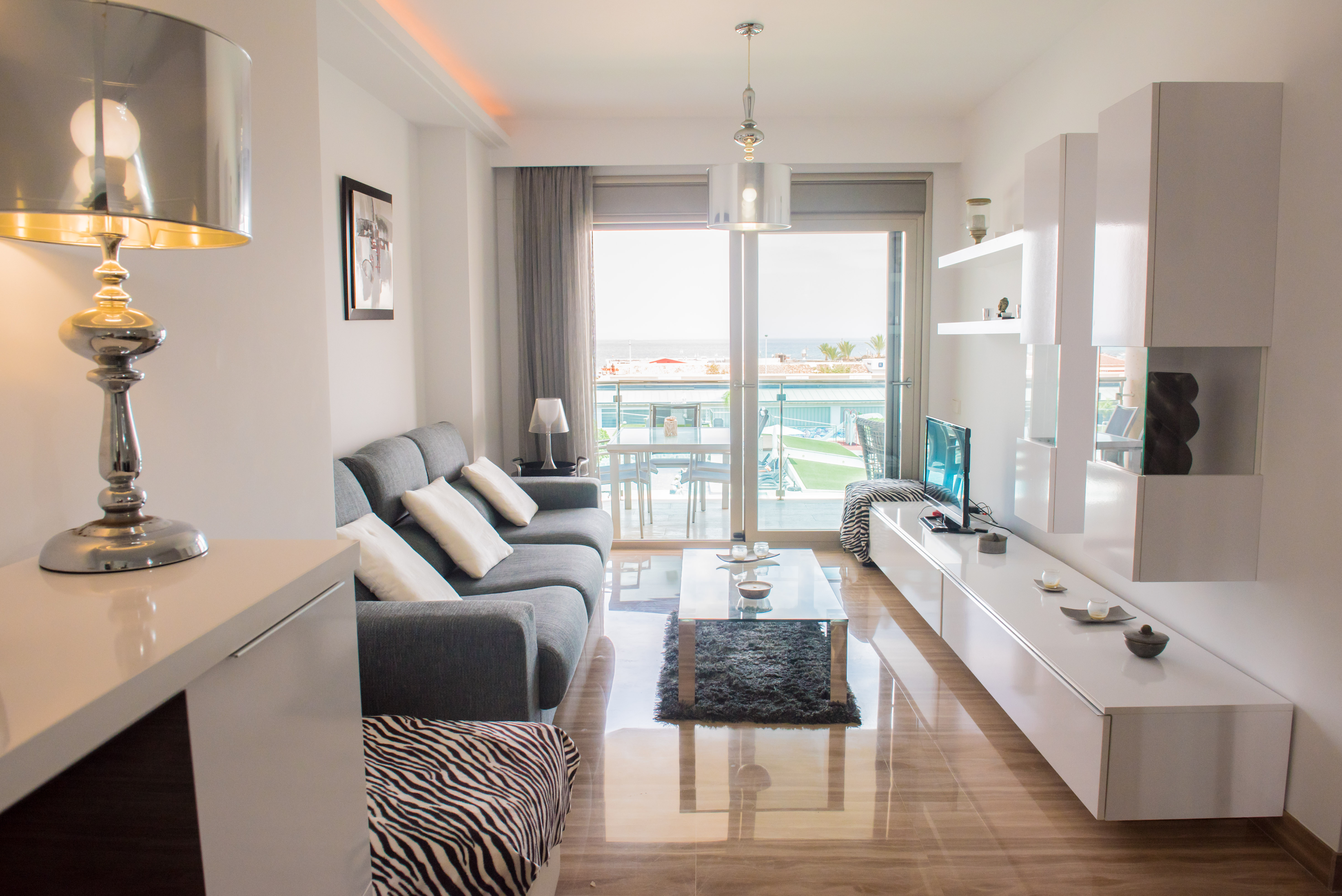 Salón moderno con suelos de mármol pulido, acceso a terraza con vistas a la piscina comunitaria y al mar. Destacan sofá gris y muebles blancos contemporáneos.