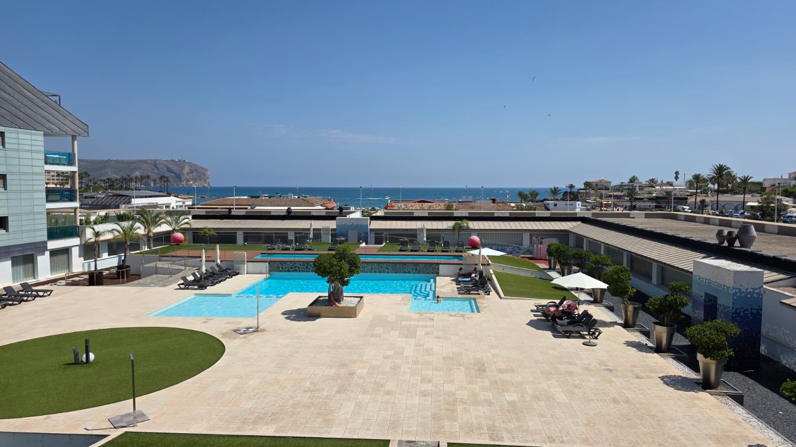 Vista aérea de la amplia zona de piscinas comunitarias con terrazas pavimentadas, césped artificial y tumbonas, en primera línea de mar con acantilado al fondo.