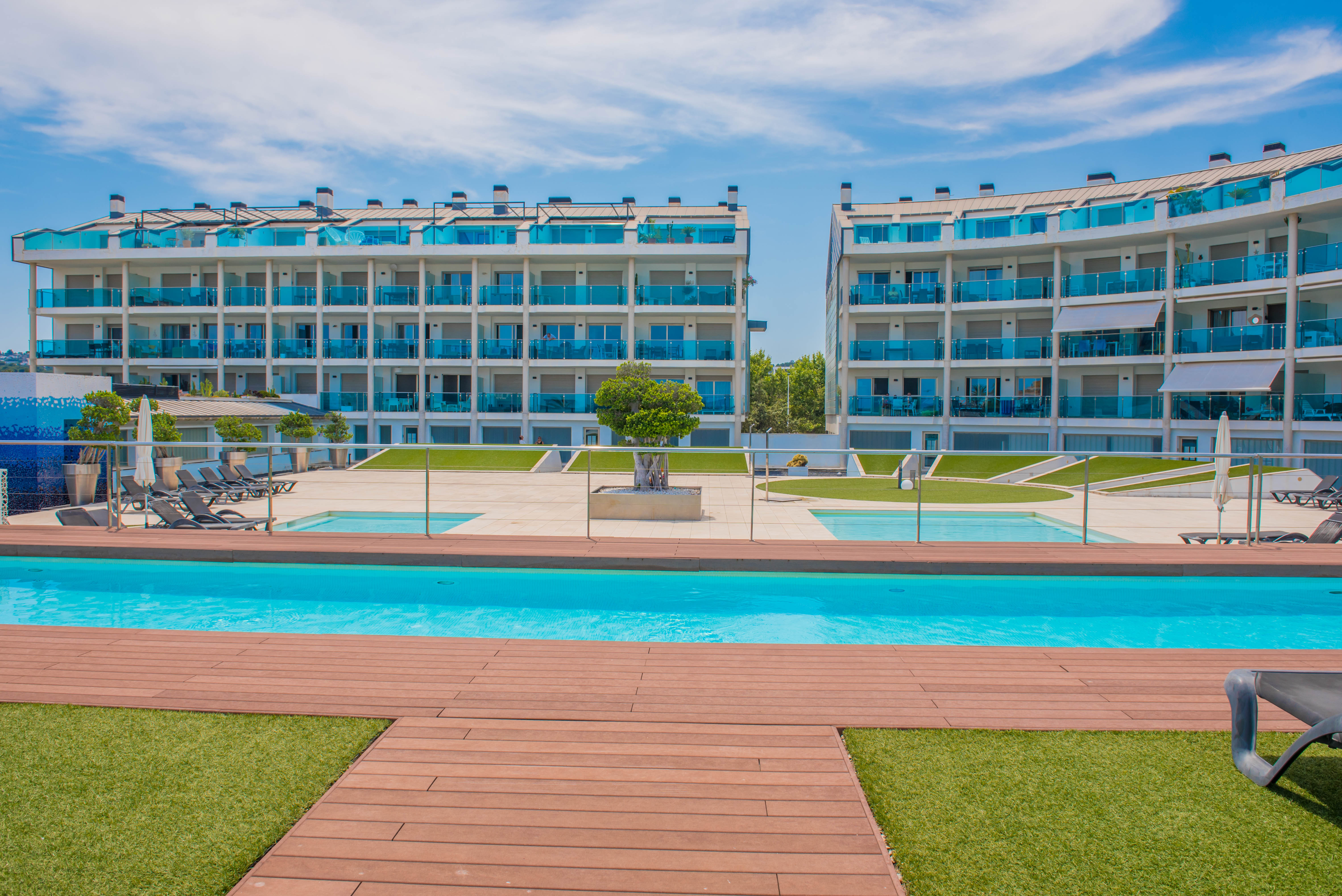 Vista de la piscina comunitaria con tarima de madera y césped artificial, flanqueada por modernos edificios residenciales con balcones de cristal azul.