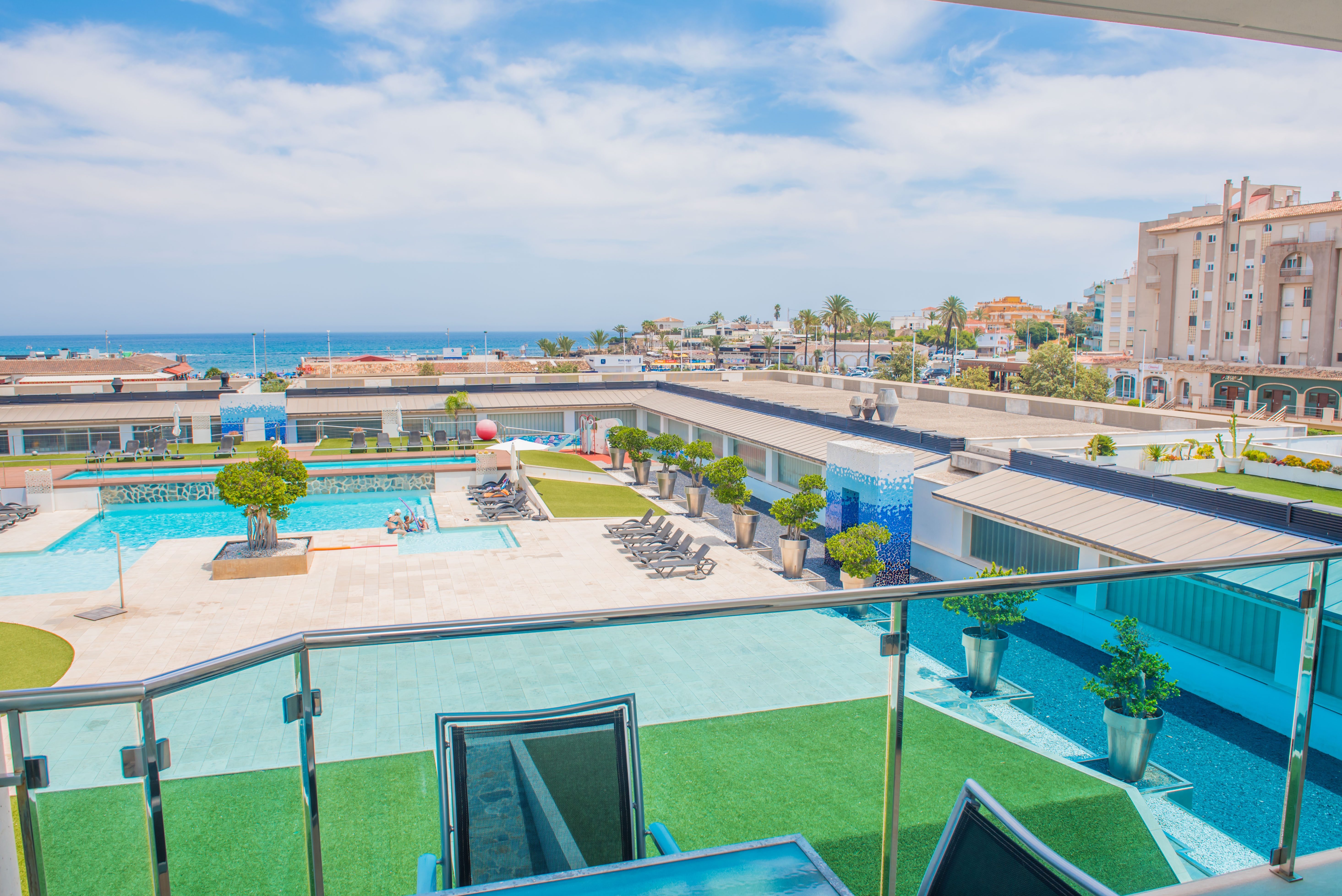 Vista elevada desde la terraza con barandilla de cristal, mostrando la piscina comunitaria, zonas de césped artificial y el mar Mediterráneo a lo lejos.