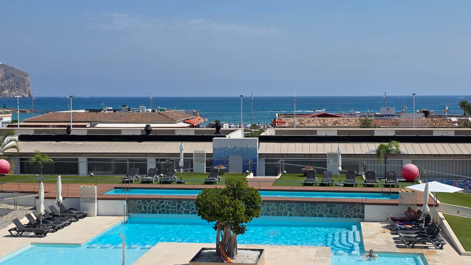 Vista panorámica de las piscinas multinivel del complejo, con tumbonas, césped artificial y vistas directas al mar Mediterráneo y la costa lejana.