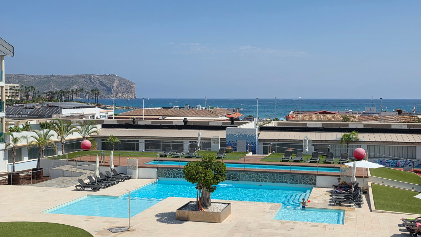 Vista panorámica del área de piscinas de varios niveles con un árbol central, rodeada de tumbonas y césped artificial, con el mar Mediterráneo y acantilados lejanos de fondo.
