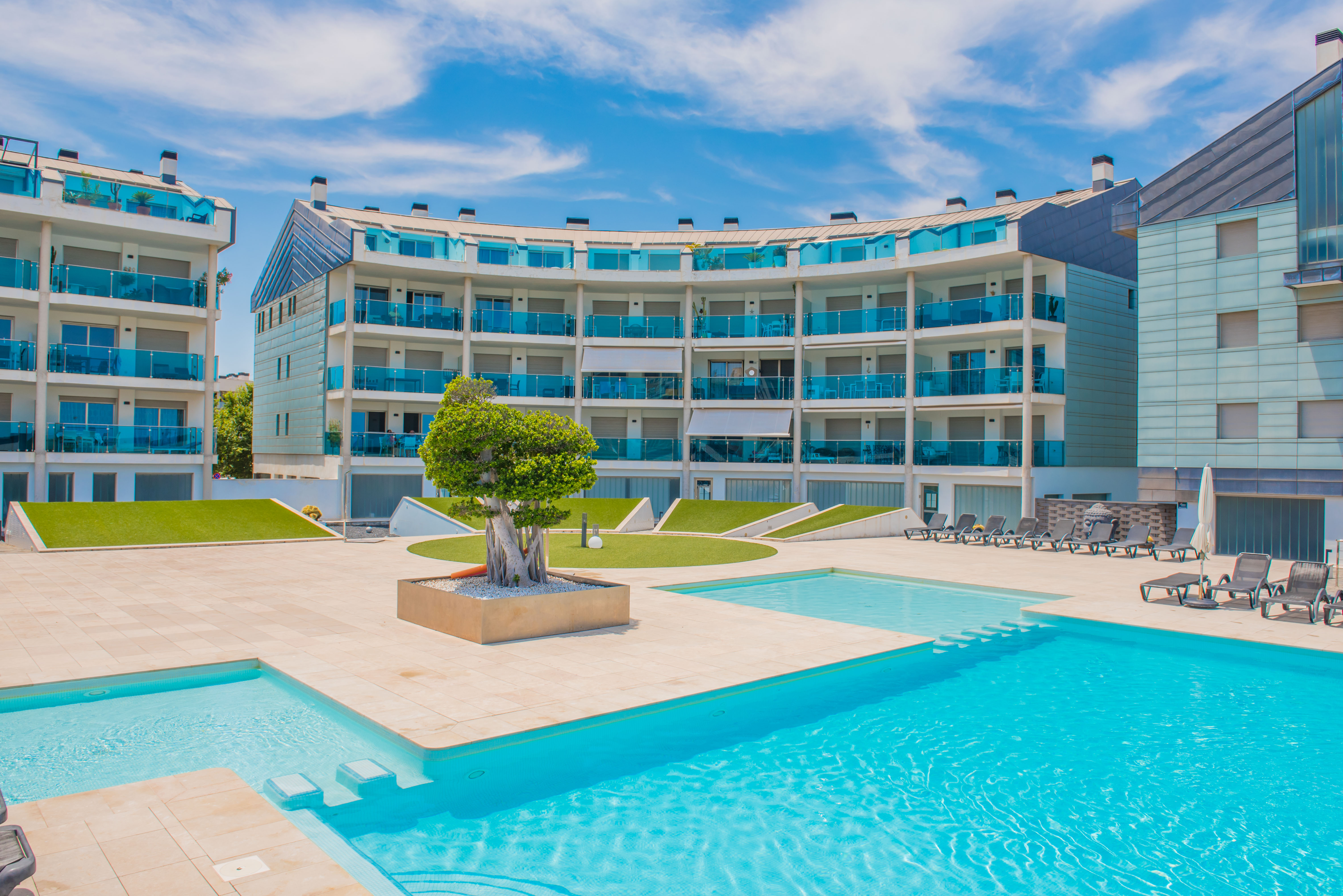 Zona de piscina comunitaria con pavimento claro, tumbonas y edificios residenciales modernos con balcones curvos de cristal azul.