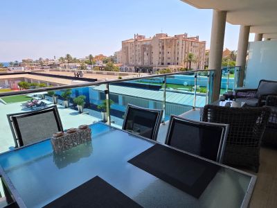 Amplia terraza con mesa de comedor de cristal y zona de estar de mimbre, ofreciendo vistas a la piscina comunitaria y al Mediterráneo bajo un cielo despejado.