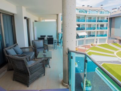 Amplia terraza cubierta con mobiliario de ratán, mesa de centro de cristal y suelo de baldosas claras. Vistas a jardines comunitarios modernos y edificio adyacente con balcones de cristal azul.