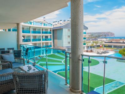 Amplia terraza privada con mobiliario de exterior de ratán y vistas directas a las piscinas comunitarias y al mar. Barandilla de cristal y suelo de gres claro.