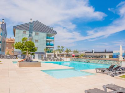 Amplia zona de piscina comunitaria con dos piscinas, pavimentación clara y numerosas tumbonas. Edificios residenciales modernos con balcones de cristal azulado enmarcan este espacio soleado.