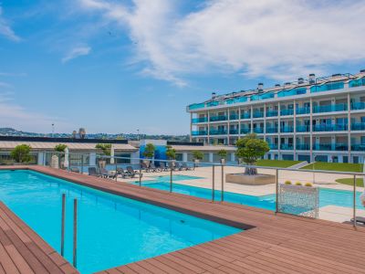 Amplia zona de piscina comunitaria con una piscina de azul brillante y una tarima de madera cálida. El moderno edificio de apartamentos, con balcones de cristal azul, domina este espacio exterior sereno.