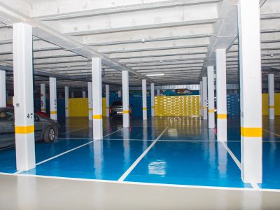 Amplio garaje subterráneo con suelo de resina azul y gris, pilares blancos con franjas amarillas y techo de hormigón visto. Se aprecian plazas de aparcamiento y paneles decorativos amarillos.