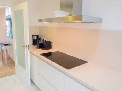 Cocina moderna con encimera clara, placa de inducción negra y campana extractora de acero inoxidable. Suelos de mármol y acceso a una sala con parquet.