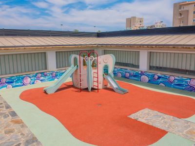 Colorida zona de juegos infantiles con estructura central de toboganes sobre superficie de caucho rojo y verde. Un vibrante mural de medusas decora la pared, bajo paneles de vidrio esmerilado.