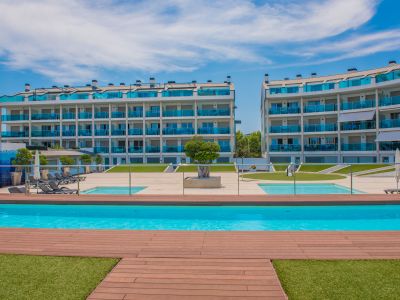 Complejo residencial moderno con dos edificios de balcones curvos de cristal azul, rodeando una gran piscina comunitaria con elegante tarima de madera y césped artificial.