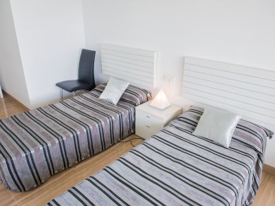Dormitorio con dos camas individuales, cabeceros blancos de lamas y ropa de cama a rayas. Una lámpara piramidal ilumina la mesita de noche entre ellas.