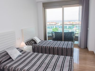 Dormitorio luminoso con dos camas individuales, ropa de cama a rayas y mesita de noche. Puertas correderas dan a un balcón con asientos exteriores y vistas parciales al mar. Suelo pulido.