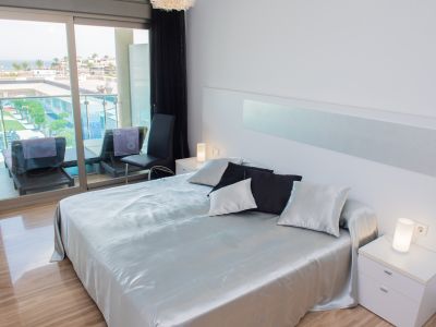 Dormitorio principal moderno con cama grande, ropa de cama satinada plateada, suelos de gres brillante y puertas correderas a la terraza con vistas al mar y piscina.