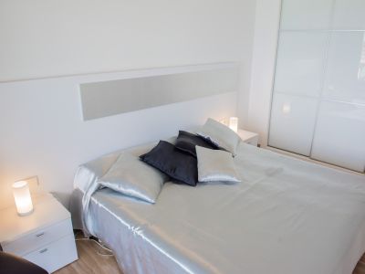 Habitación moderna con cama de sábanas satinadas, cabecero blanco y mesitas de noche con lámparas cilíndricas. Armario empotrado blanco con puertas correderas de cristal esmerilado.